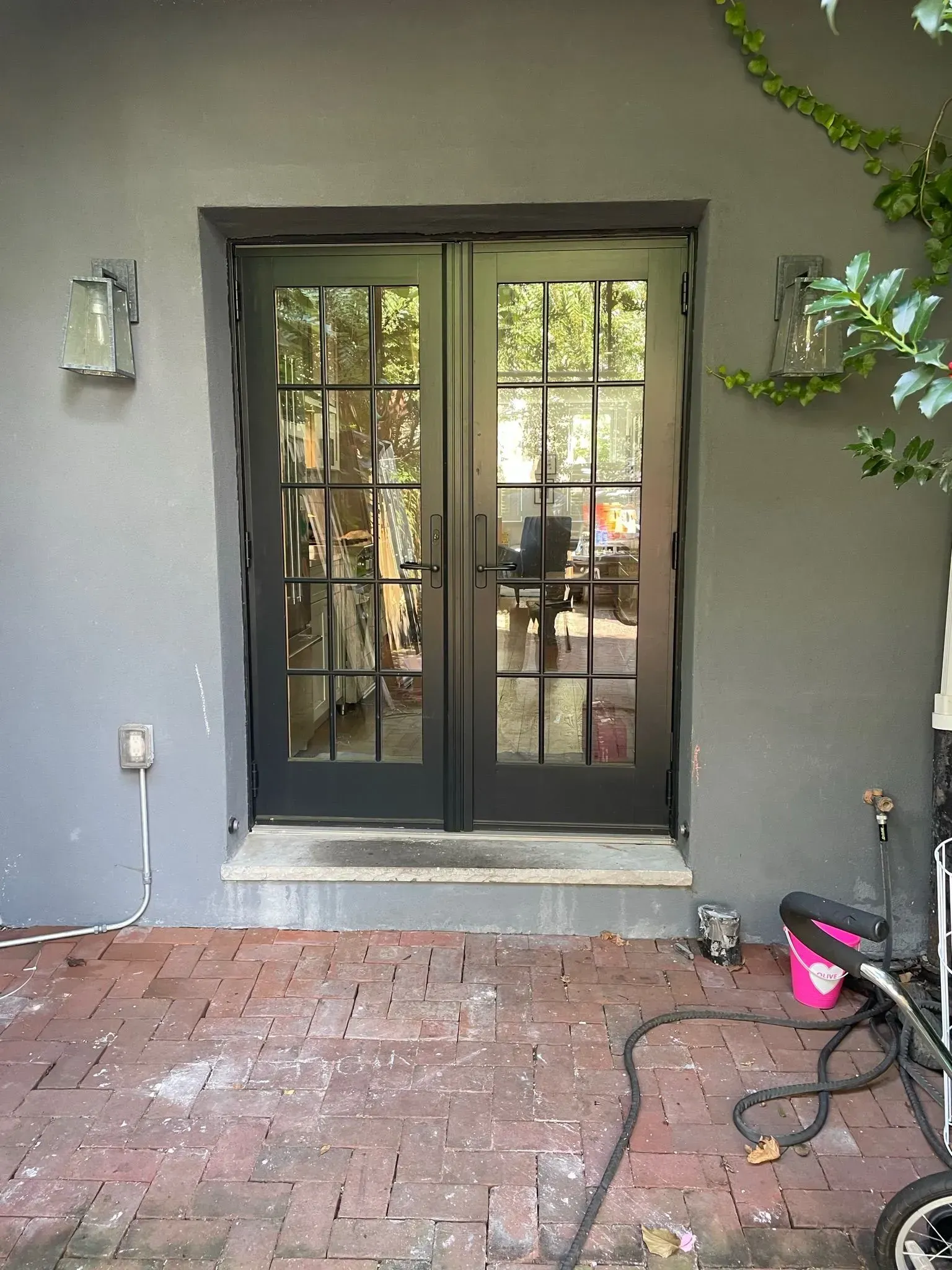 Black French doors on a gray wall with brick patio; sconces and greenery are flanking.