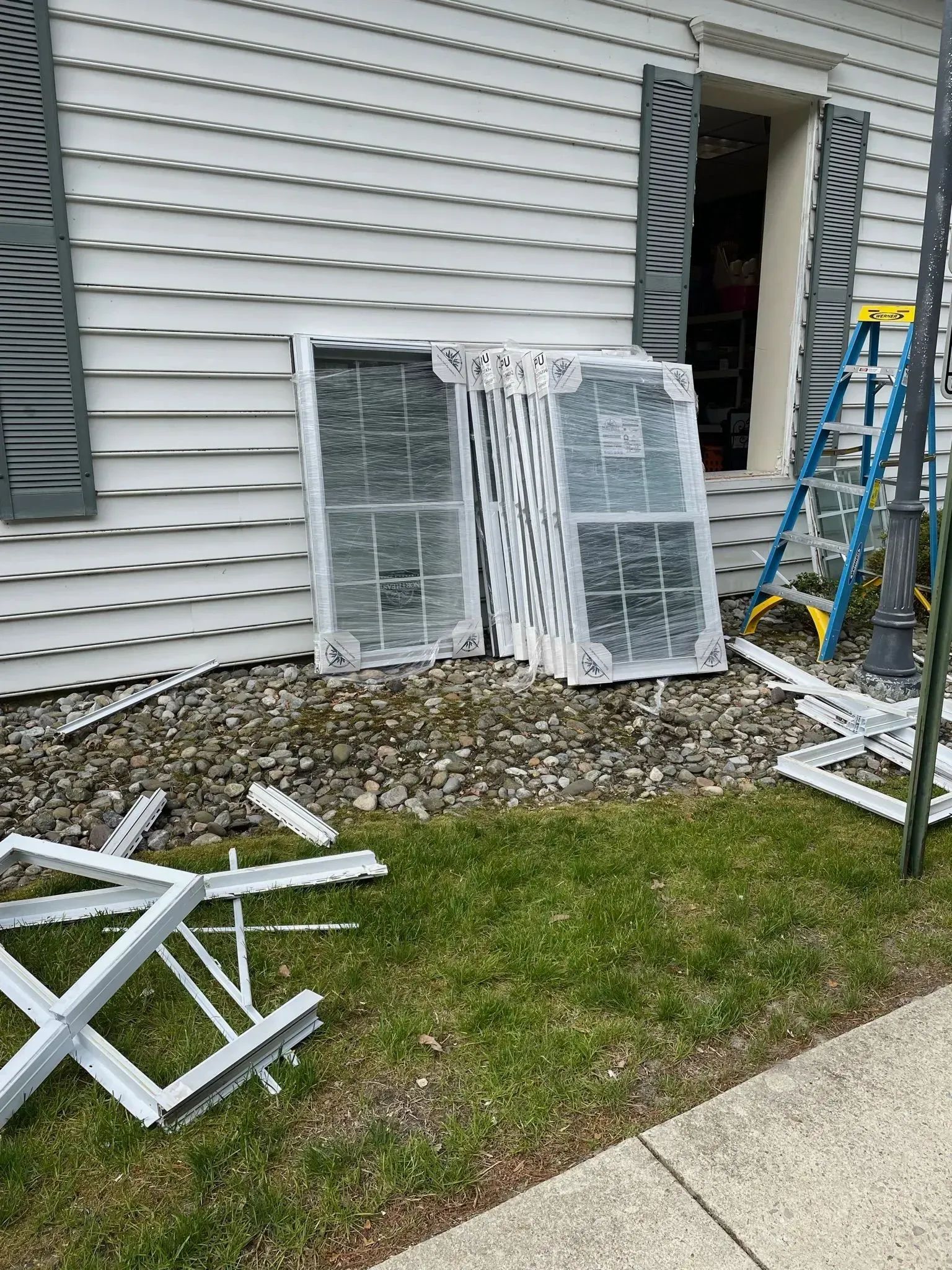 New windows wrapped in plastic, lying on the ground next to a house with shutters; a ladder stands nearby.