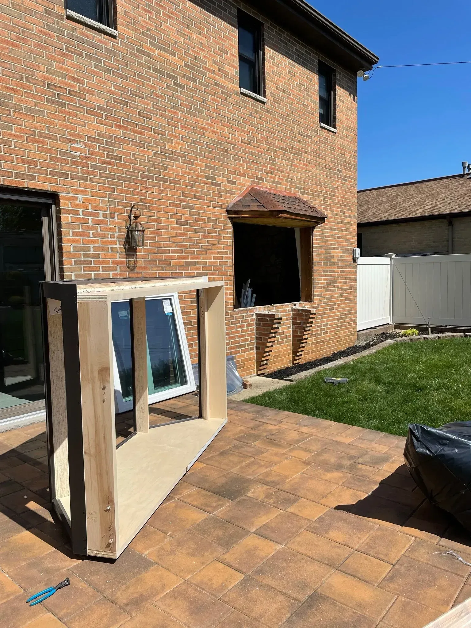 A rectangular wooden frame with a window leans on a brick patio in front of a brick building.