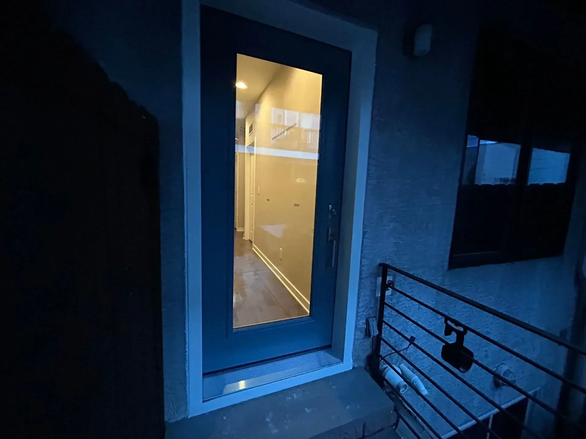 Exterior view of a dark doorway with a glass panel, leading to a brightly lit hallway.