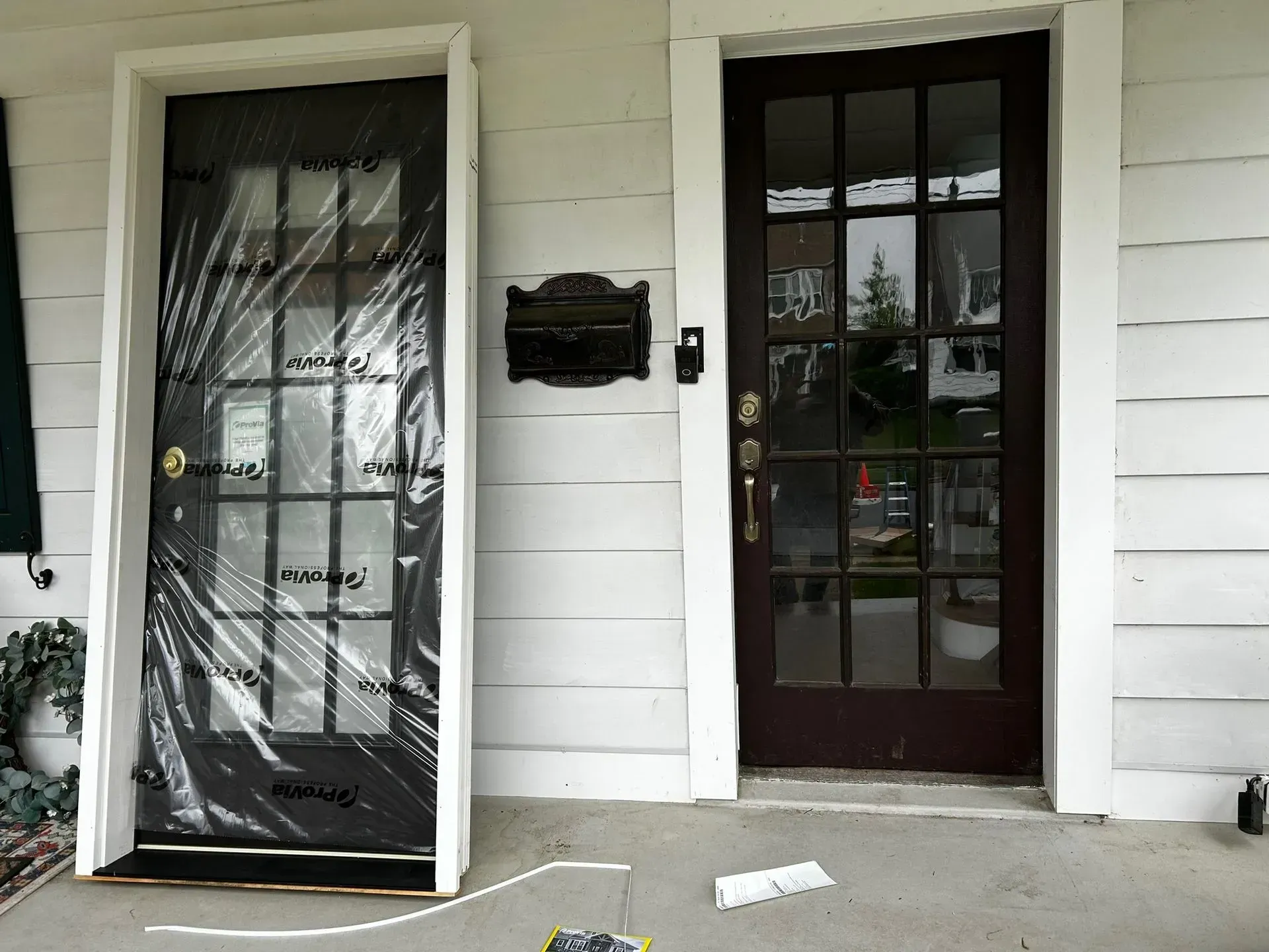 Two dark doors with white trim on a white siding house. A screen door is wrapped in plastic.