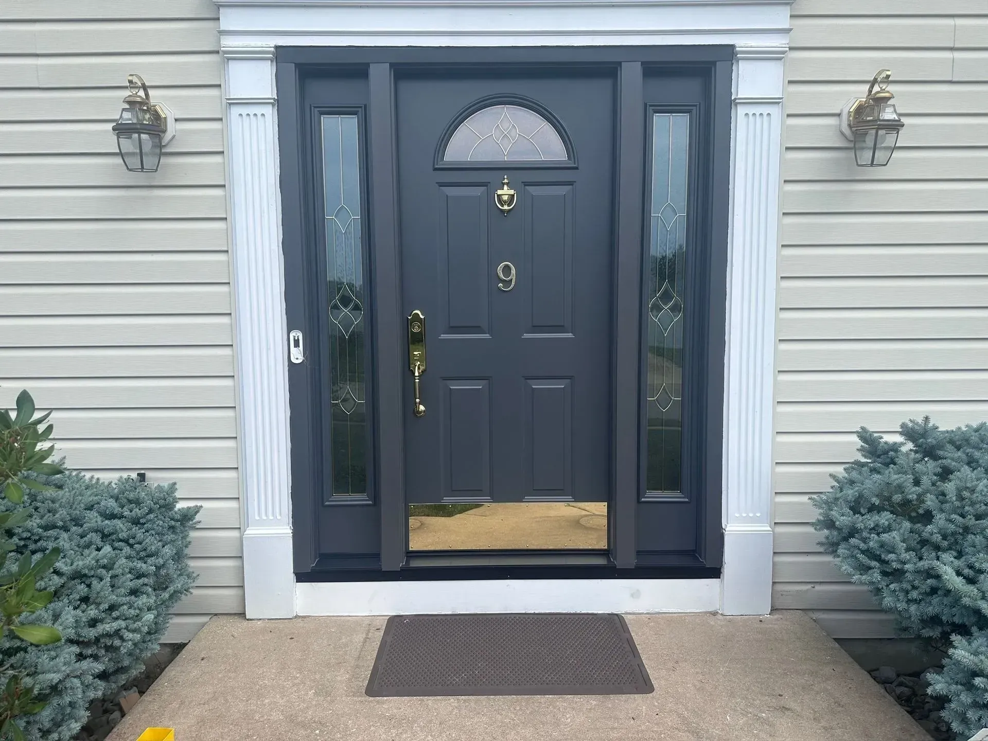Gray front door with sidelights and gold hardware. Exterior of house with bushes and lights.
