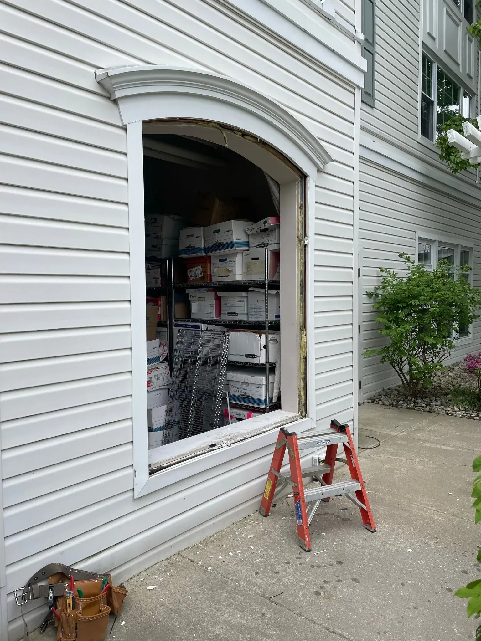 Window opening in a white building, revealing shelves of boxes; small red ladder in front.