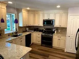 Cream-colored kitchen cabinets and appliances with a granite countertop, wooden floor, and a window.