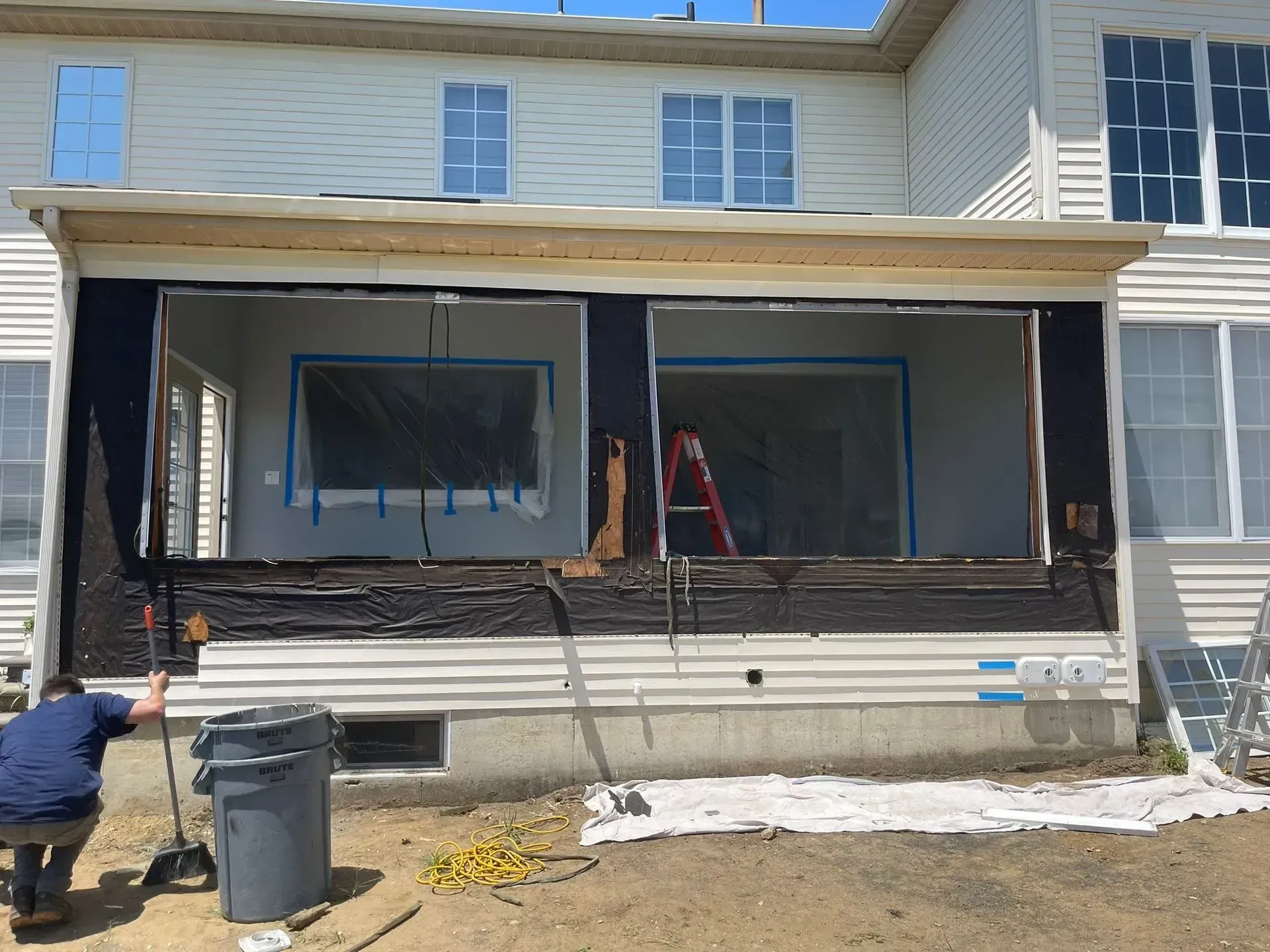 Exterior of a house under renovation.  Worker on the left. Windows are framed and taped.  Black siding is exposed.  Red ladder inside.