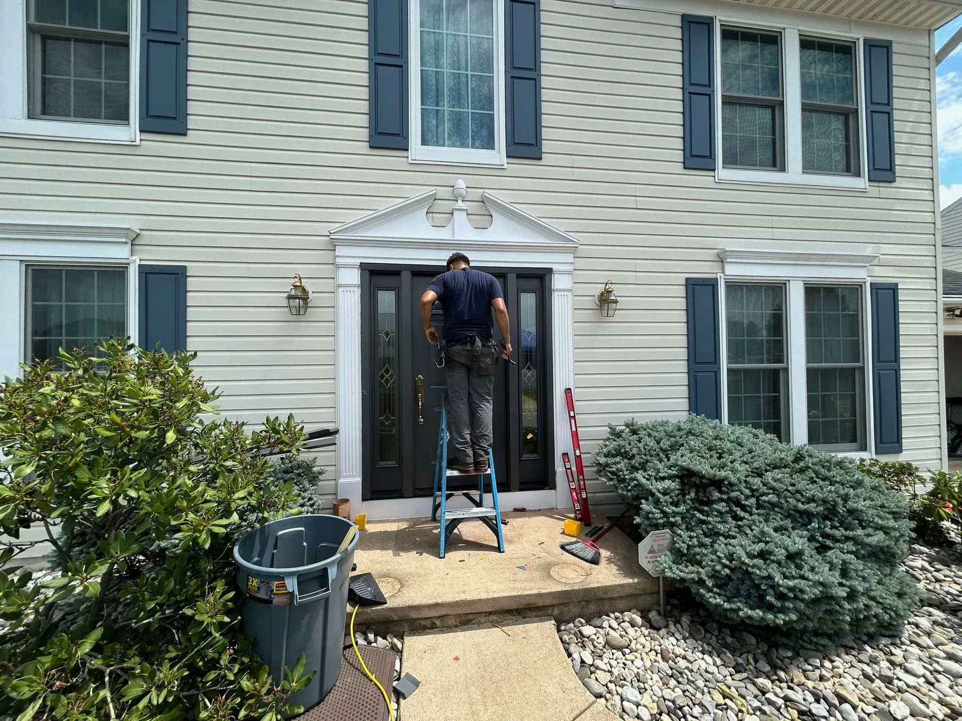 Person on a step ladder working on a front door. Beige house with blue shutters; bushes and trash bin in foreground.