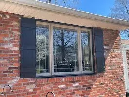 A brick house with a window framed by black shutters. The window reflects a tree.