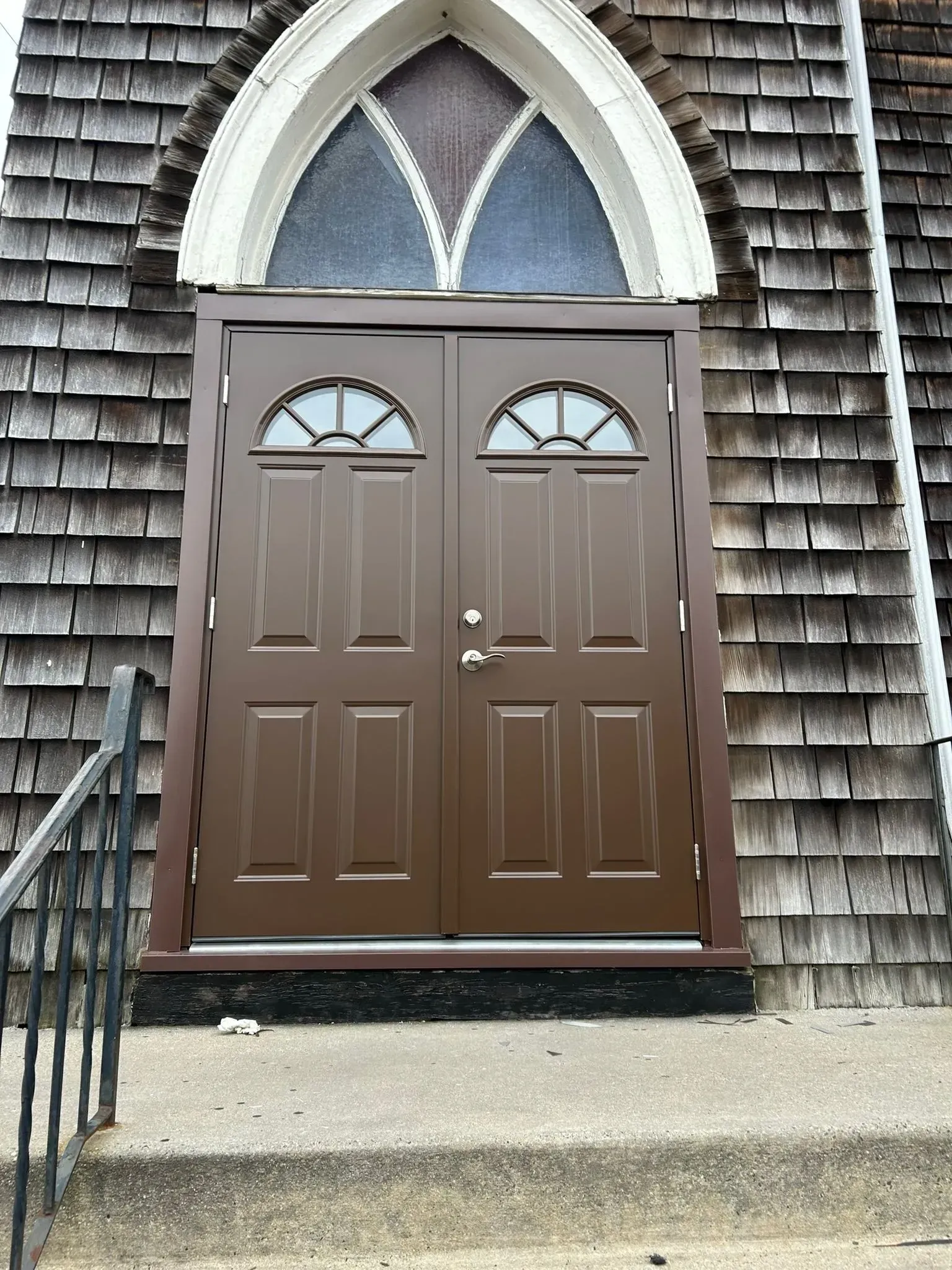 Brown double doors with arched transom window, on building with cedar shake siding.
