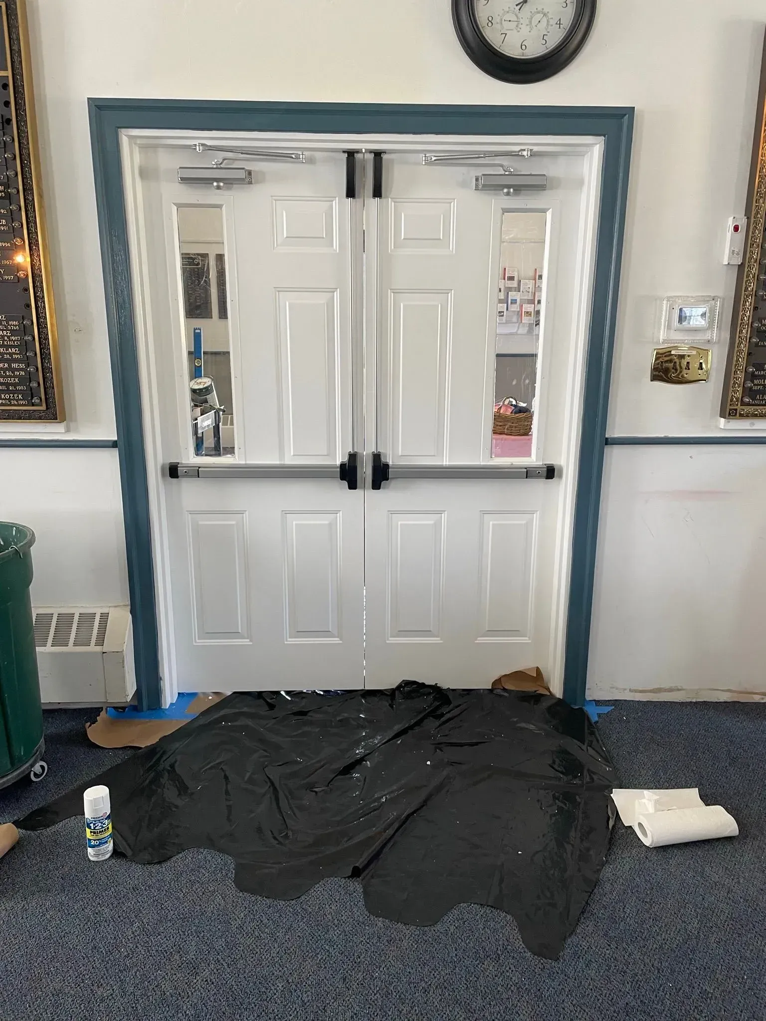 Double white doors with glass panels and safety bars, framed in blue, against a wall. A black tarp and supplies are on the floor.