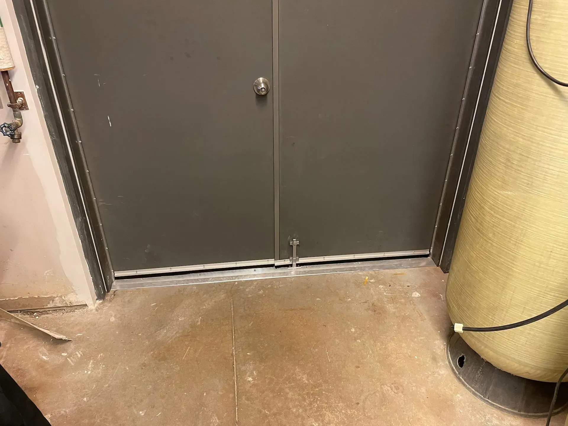 A gray double door with a silver threshold, partially open, against a brown floor.