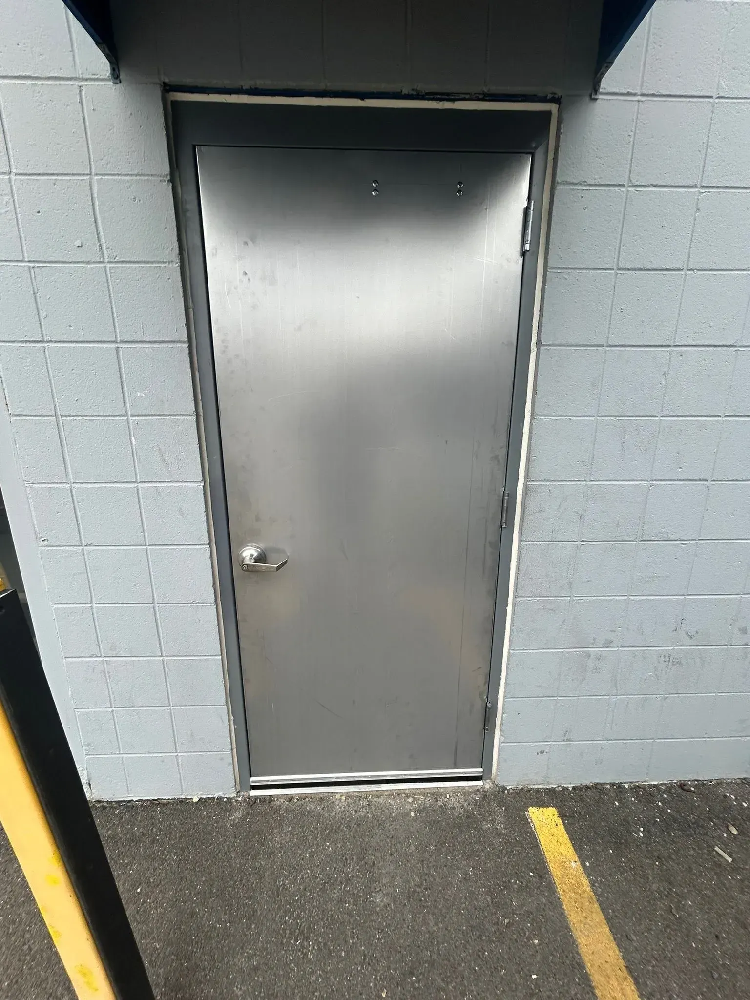 Silver metal door set in a concrete block wall, with a small handle and a parking area at the bottom.