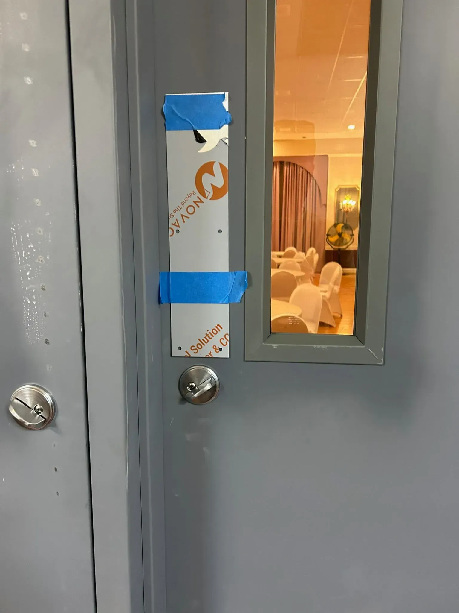 Gray door with a rectangular window. A metal plate is taped to the door, covering an area.