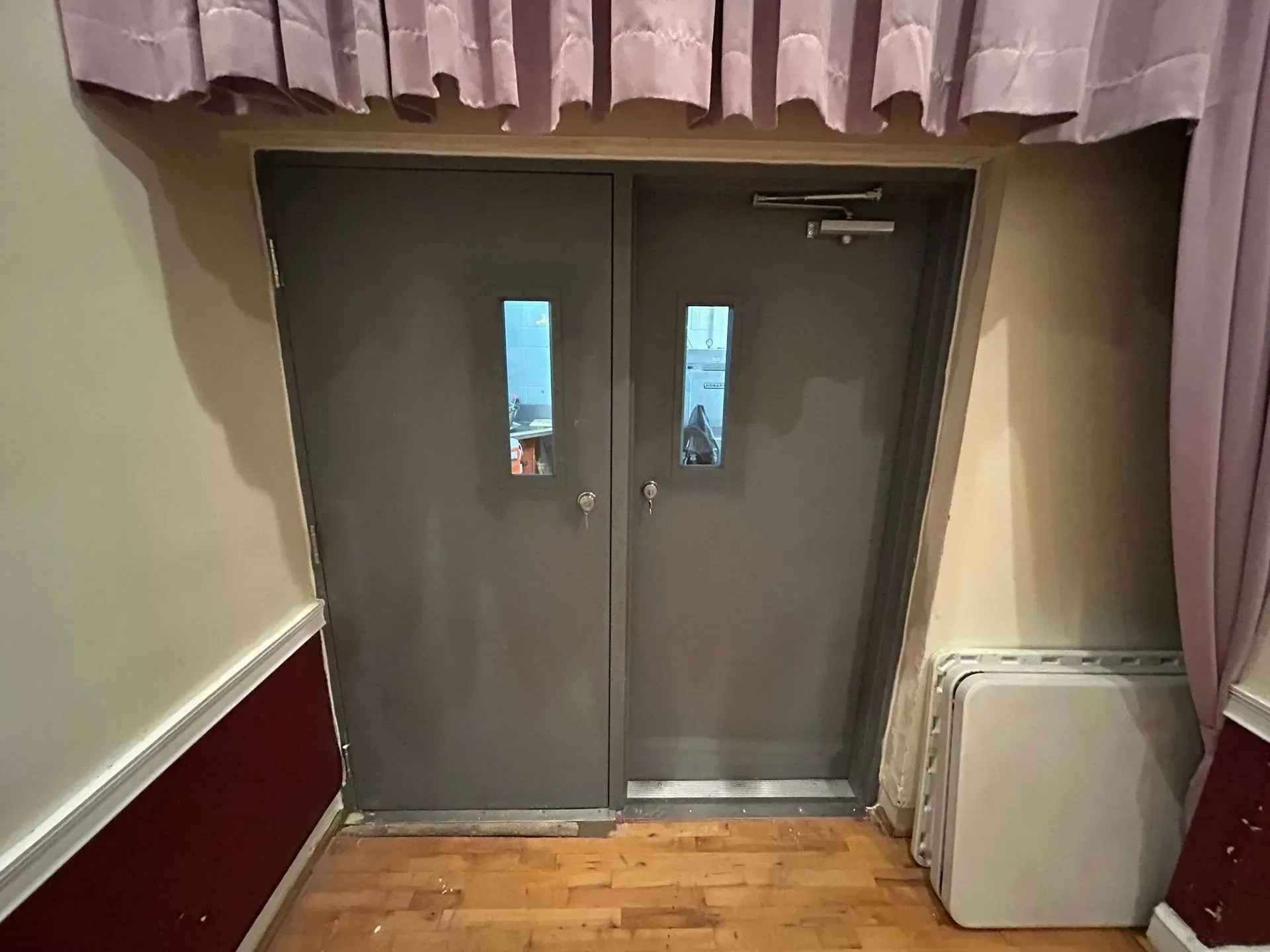 Gray double doors with small windows, flanked by maroon and beige walls. Curtains hang above.