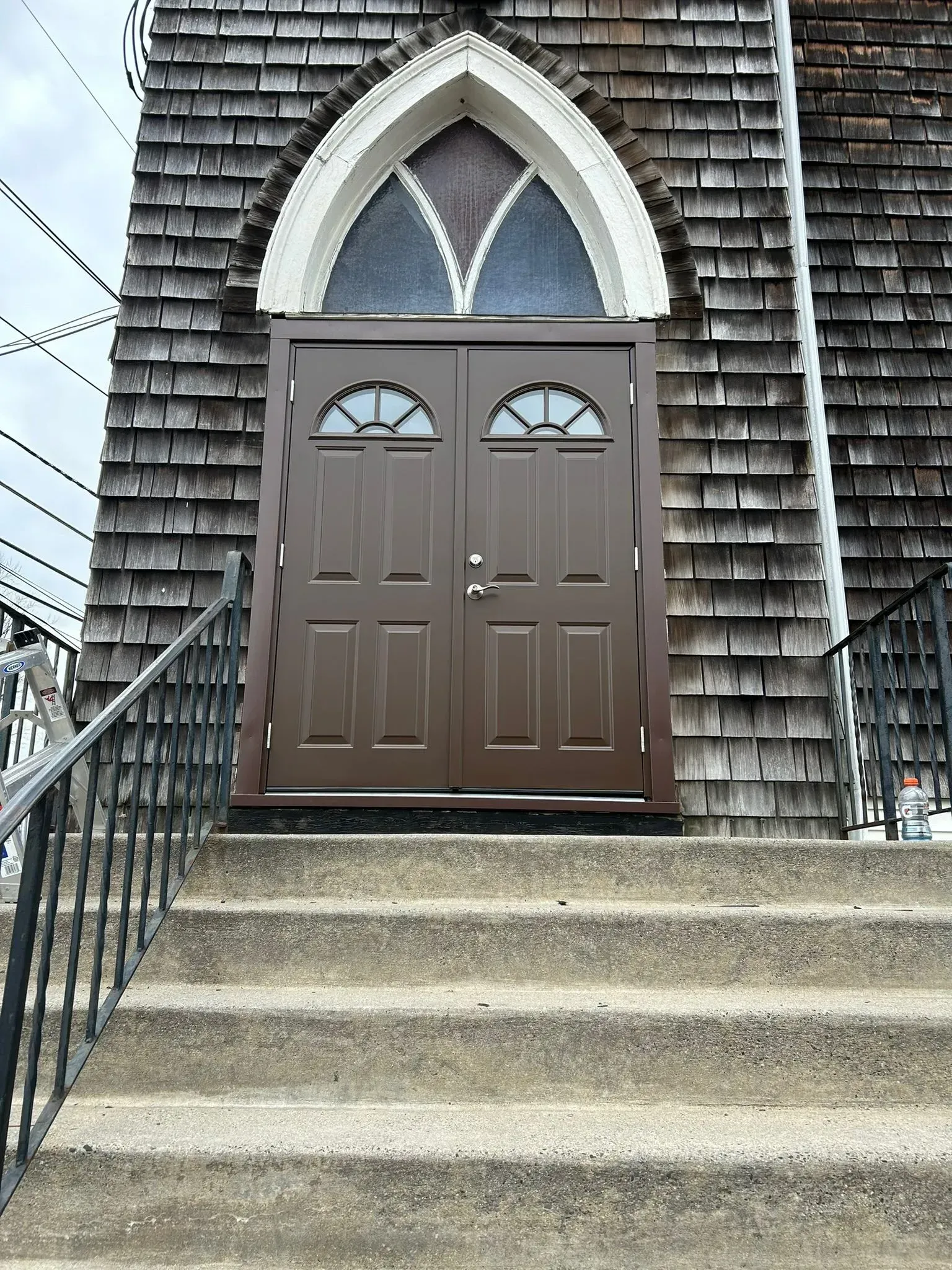 Brown double doors with arched window above, set into a building with wood shingle siding; concrete stairs with black railing lead up to the entrance.