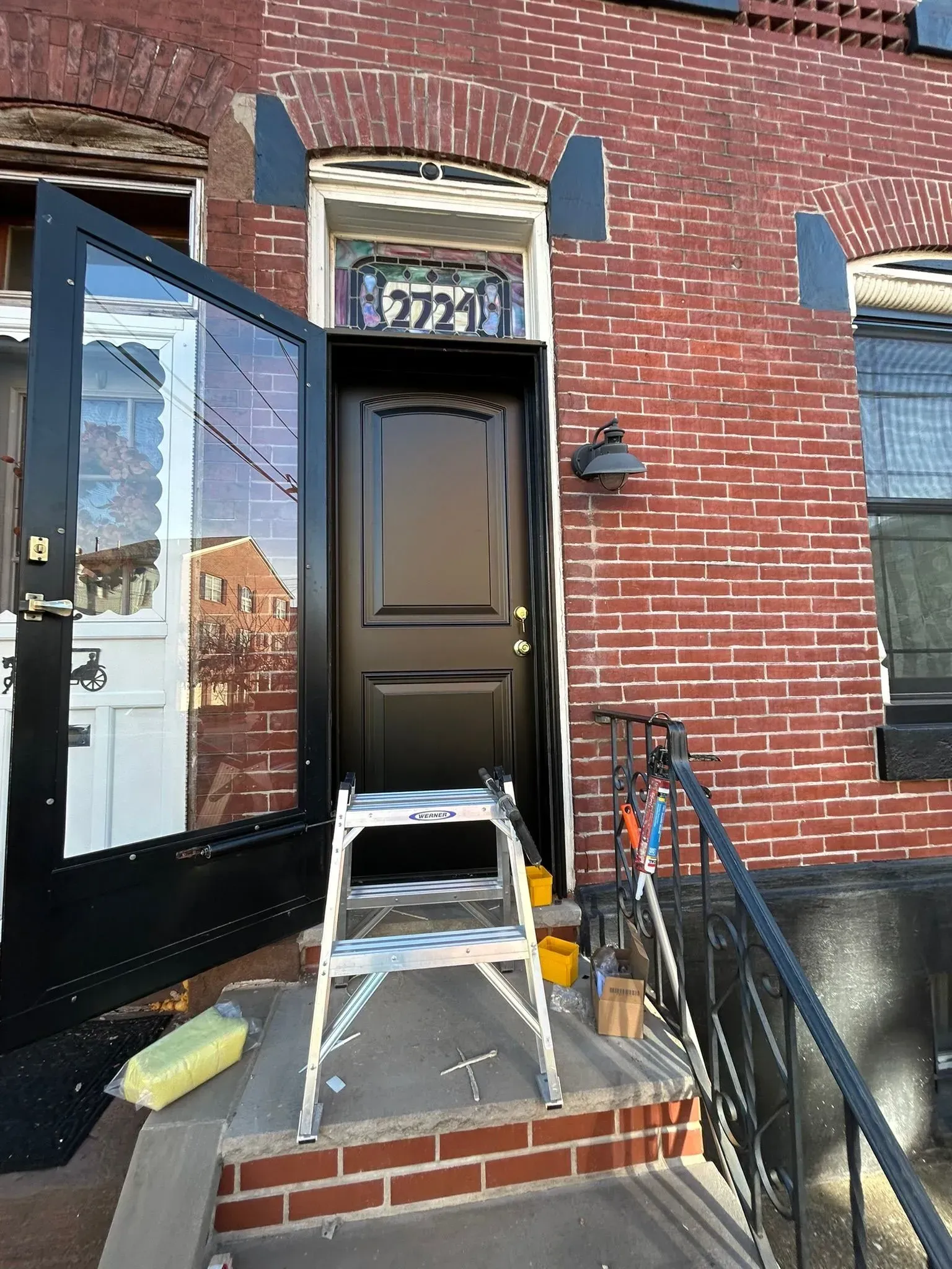 Exterior view of a brick building with a black front door open. A stepladder stands in front.