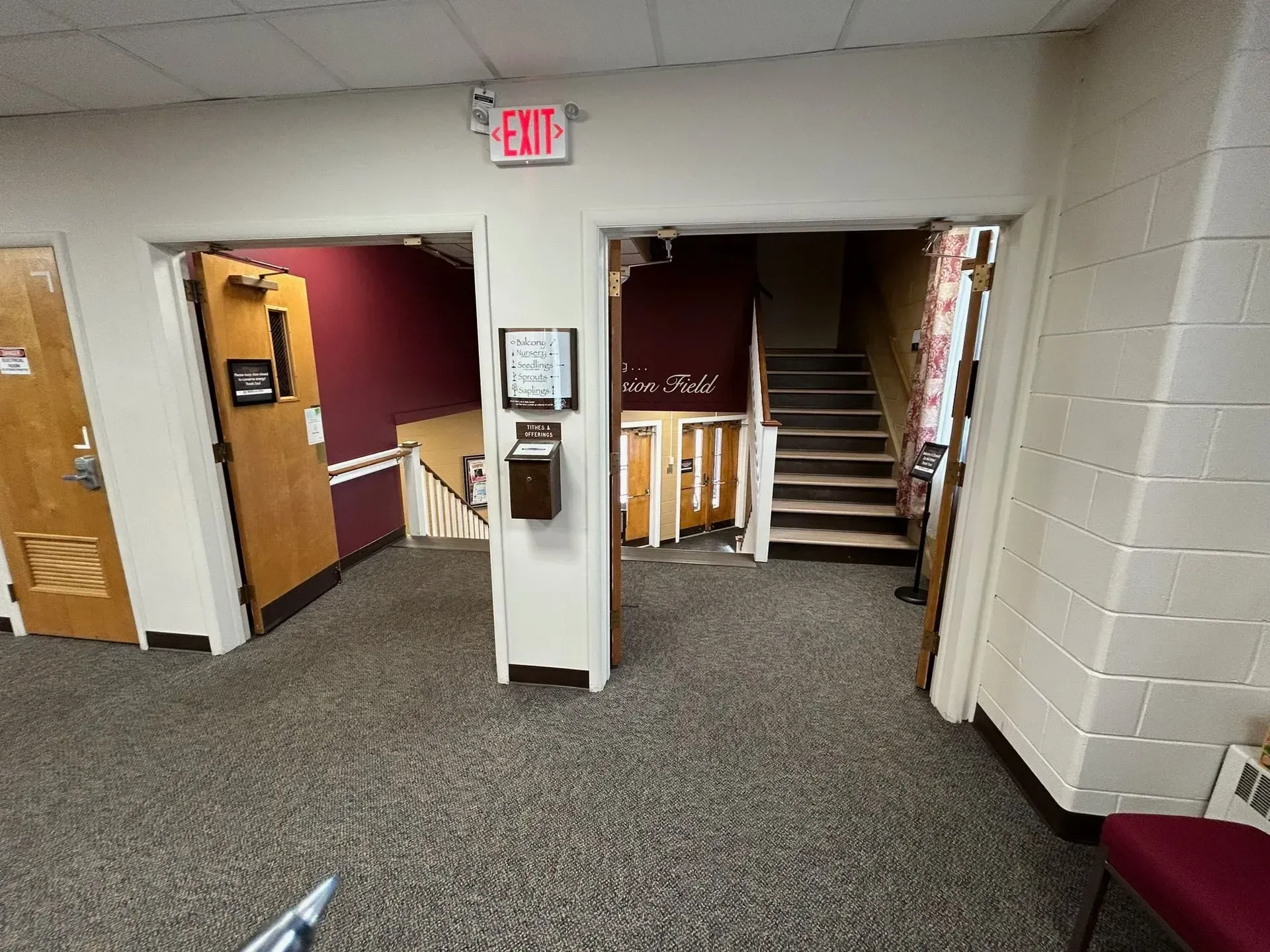 Hallway with doors, stairs, and an exit sign. Dark red and brown walls, beige trim, and gray carpet.