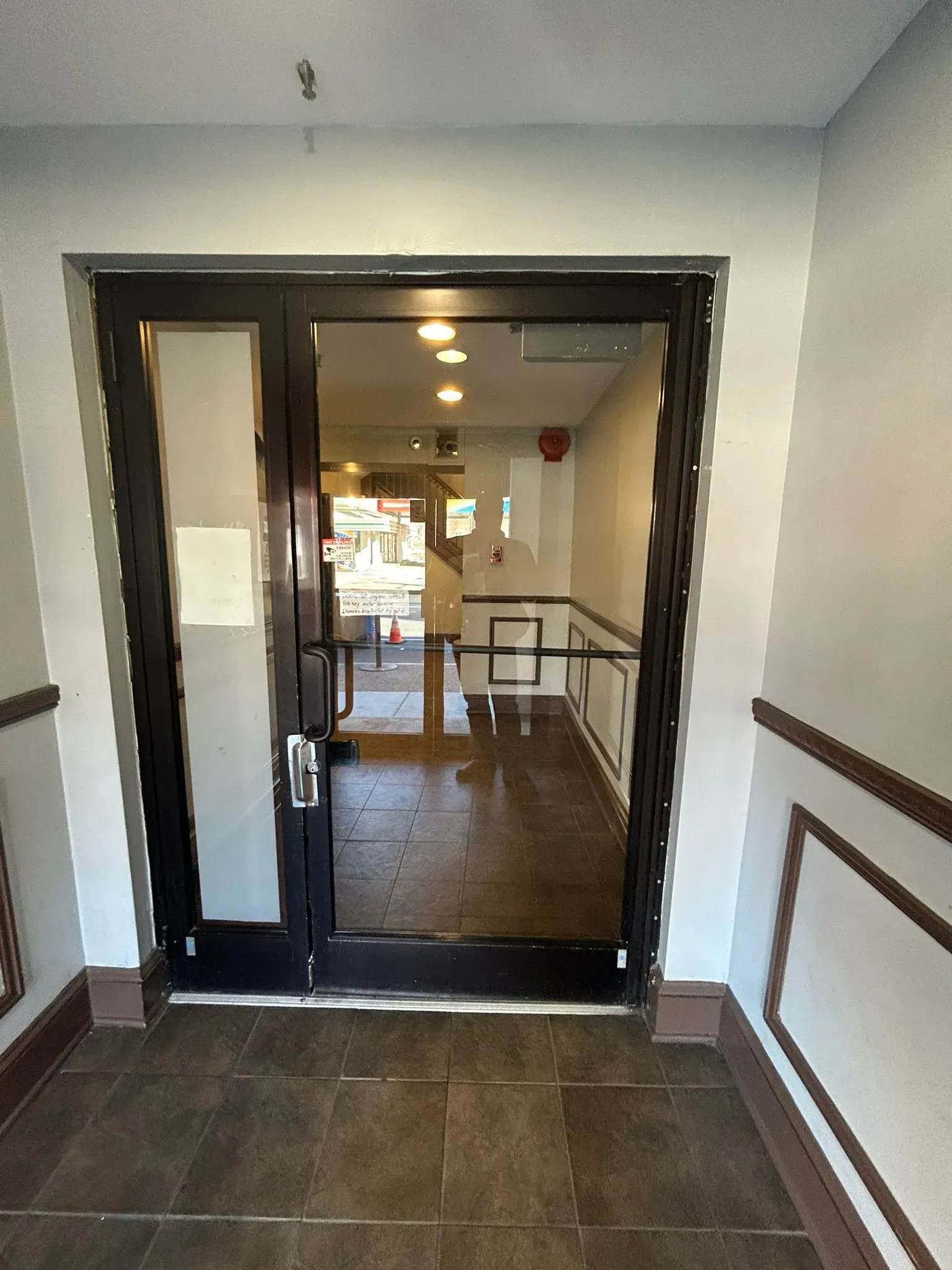 Black framed glass door leading into a building. Brown tiled floor and white walls.