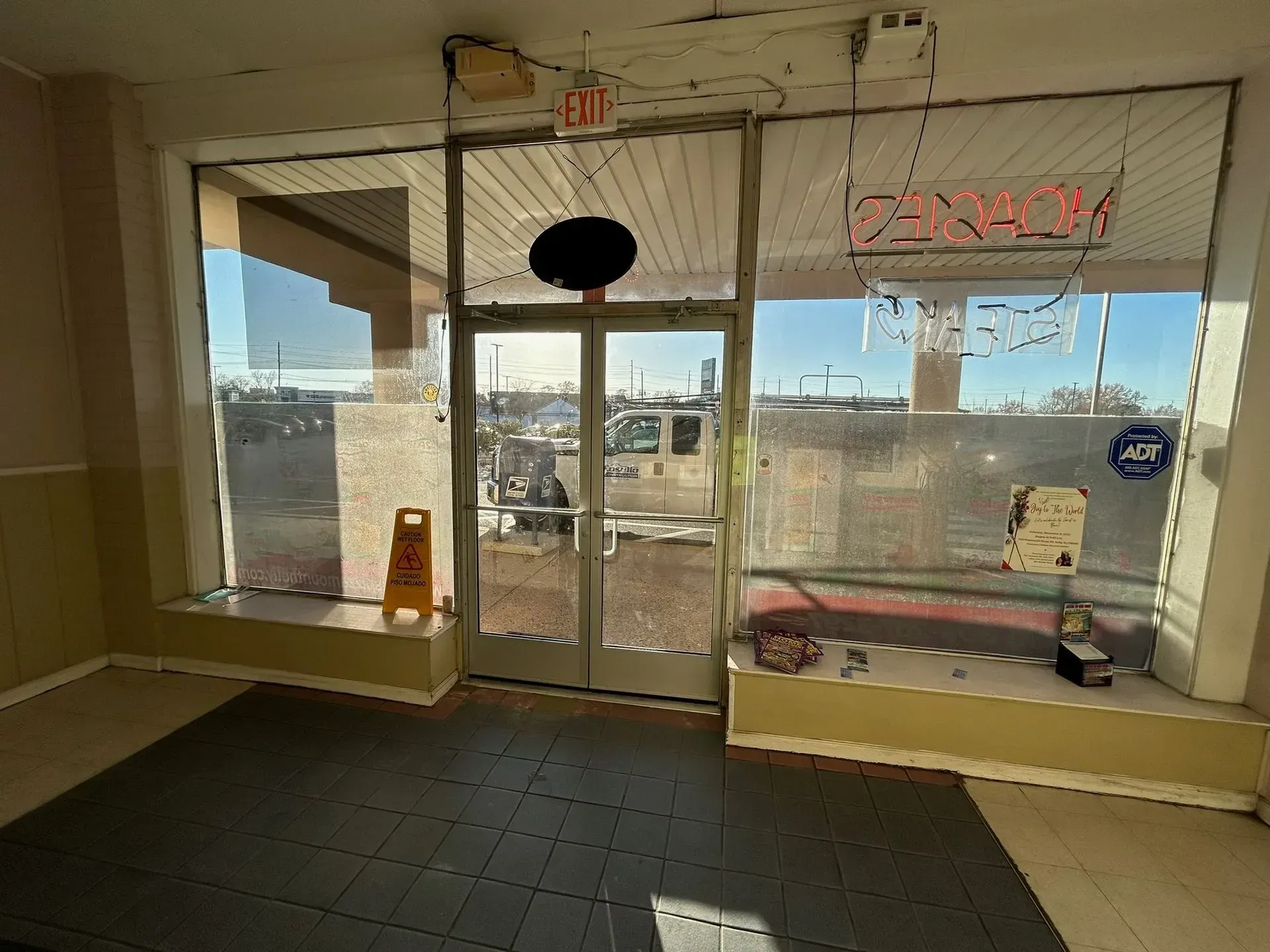 Empty storefront with a glass door and window. Yellow warning sign, neon sign visible.