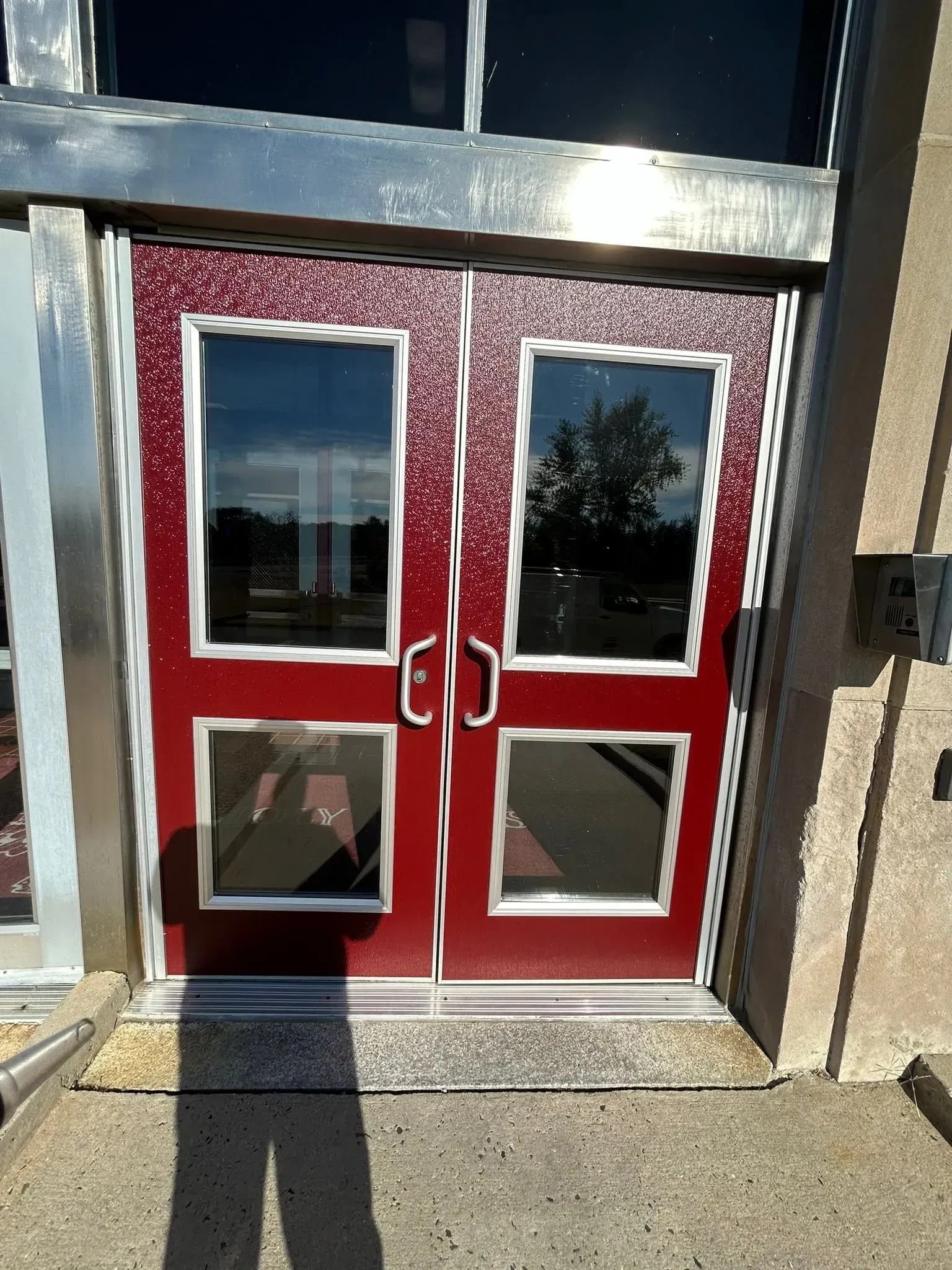 Red double doors with rectangular windows, in a building entrance. Silver trim and a concrete surface.