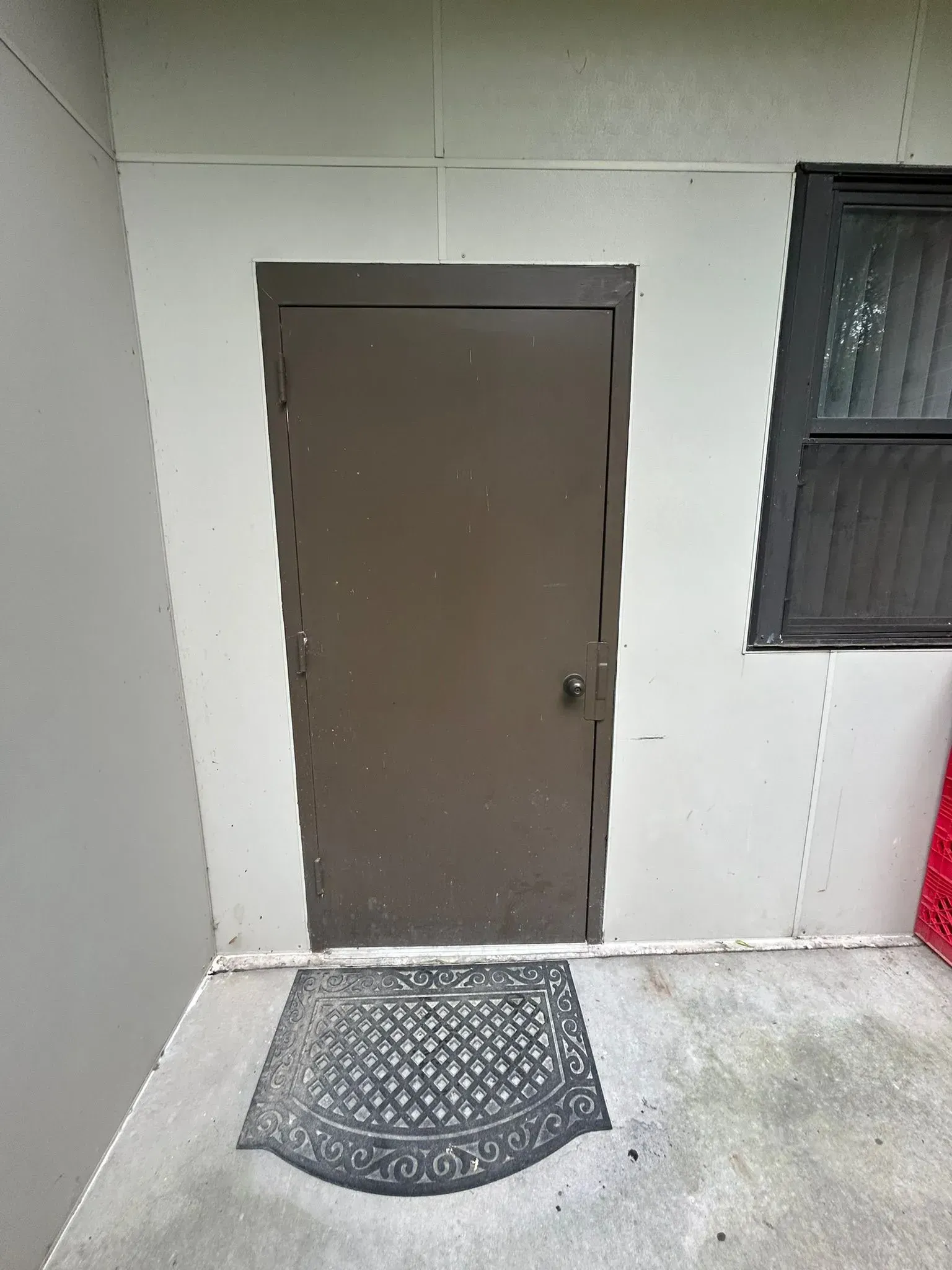 Brown metal door set in wall, with mat. Right, a window with closed blinds.