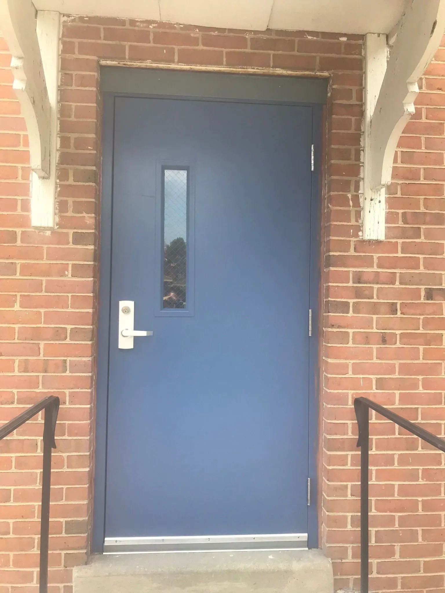 Blue door with rectangular window set in a brick wall. Silver handle and railing present.