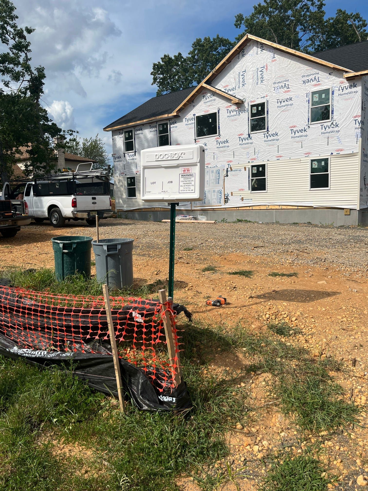 Construction site with two-story house wrapped in Tyvek. White truck, trash cans, and safety fence are in the foreground.