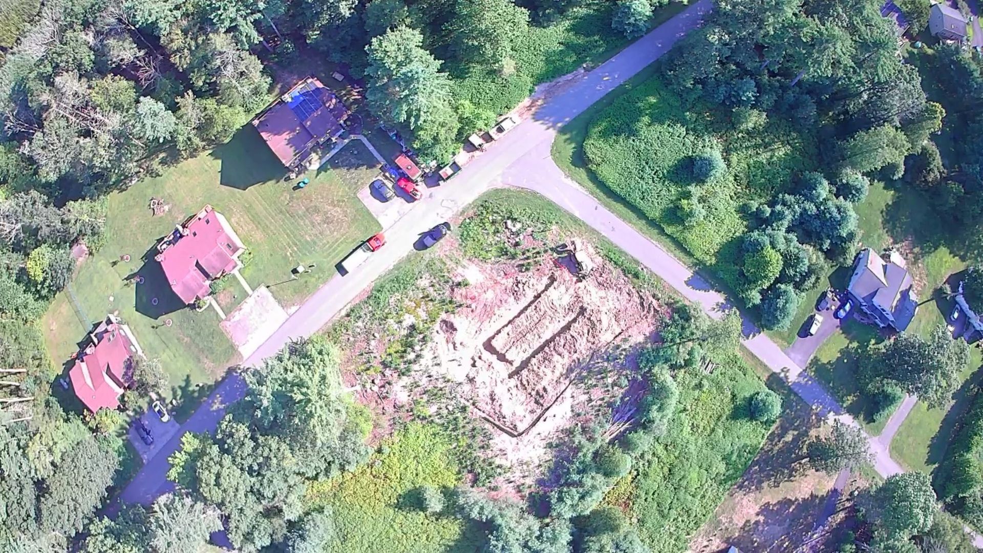 Aerial view of a cleared construction site at a road intersection, surrounded by houses and greenery.