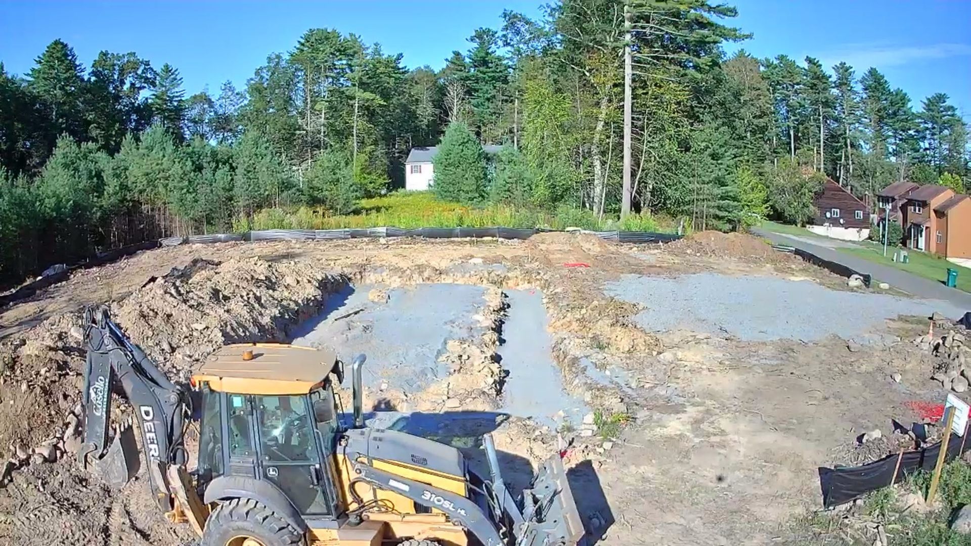 Yellow and gray construction vehicle excavating a lot, trees and a house in the background.