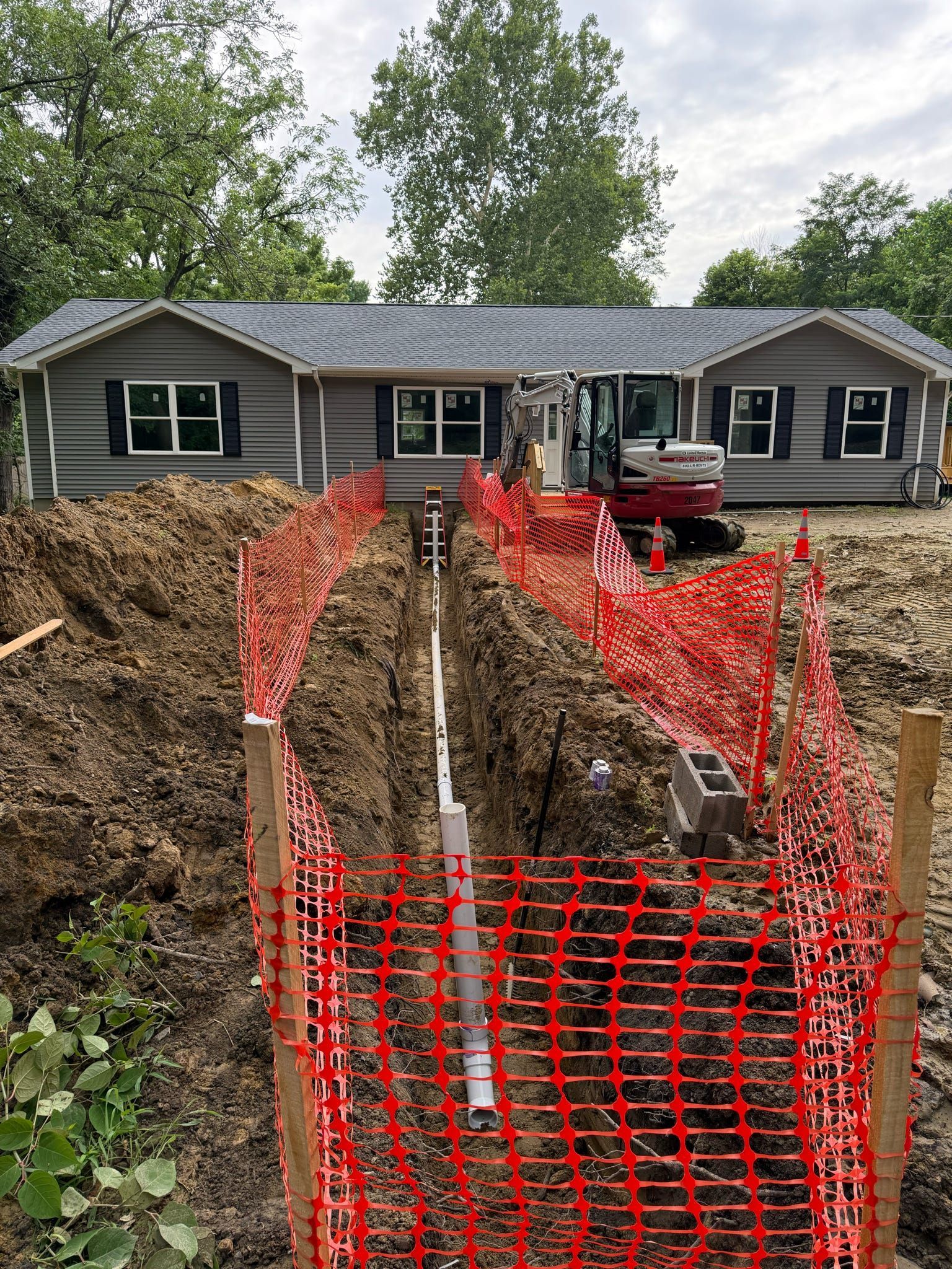 Trench with orange safety fence in front of a gray house during construction, small excavator in the background.