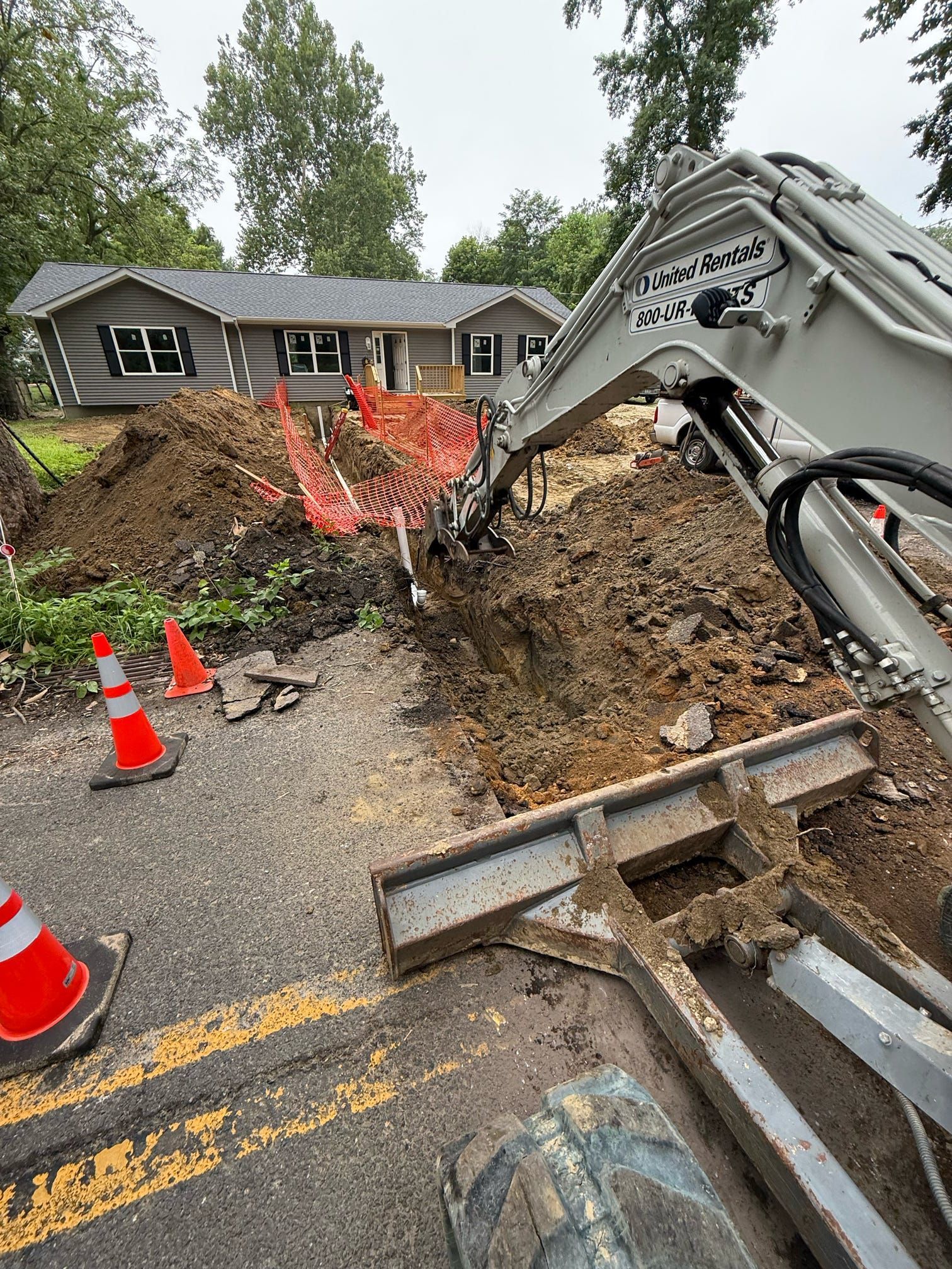 Excavator digging a trench near a house, with safety cones on the asphalt road and orange fencing.