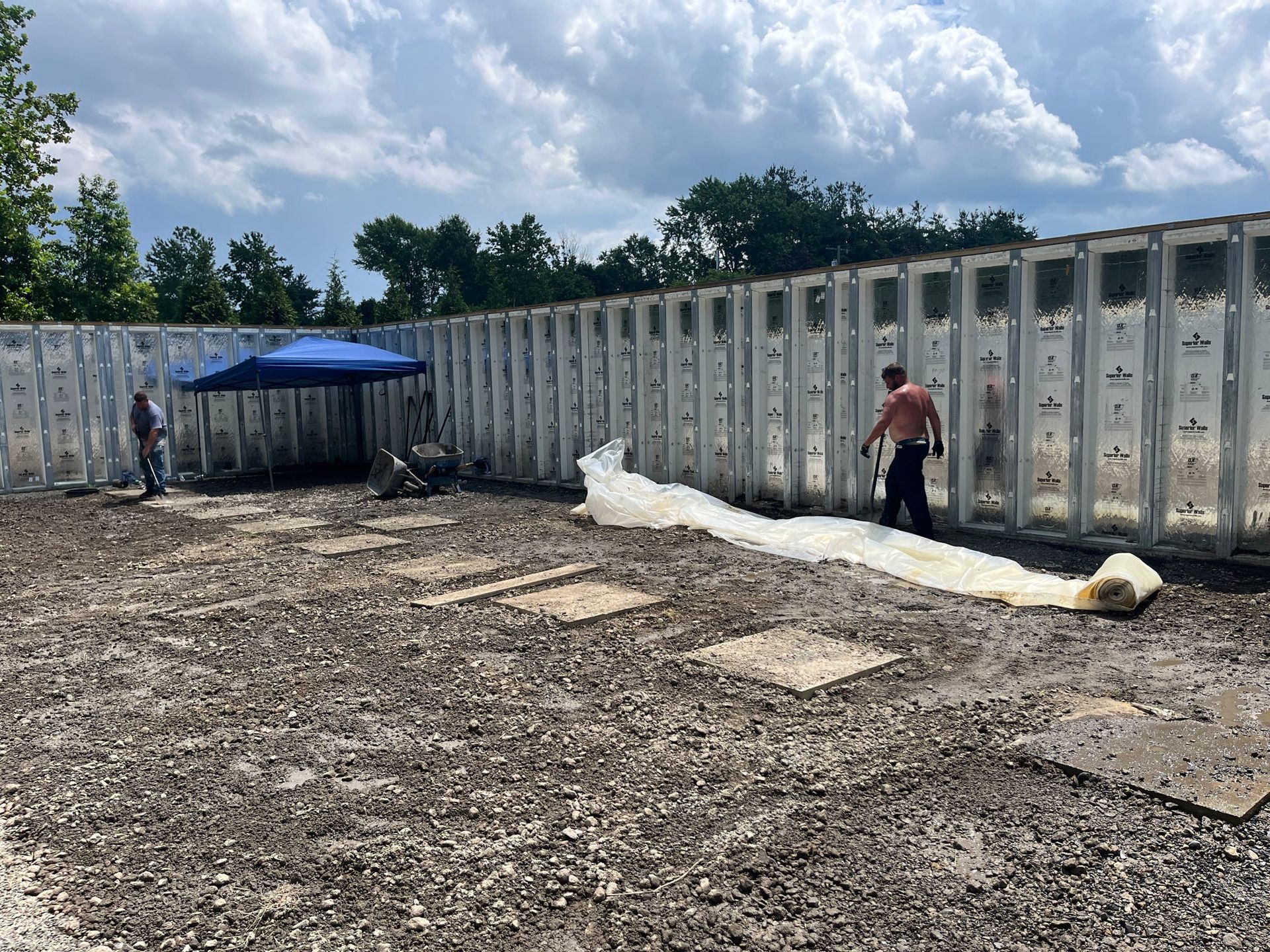 Workers roll out white material on a gravel surface next to a tall, segmented wall. A blue canopy provides shade.