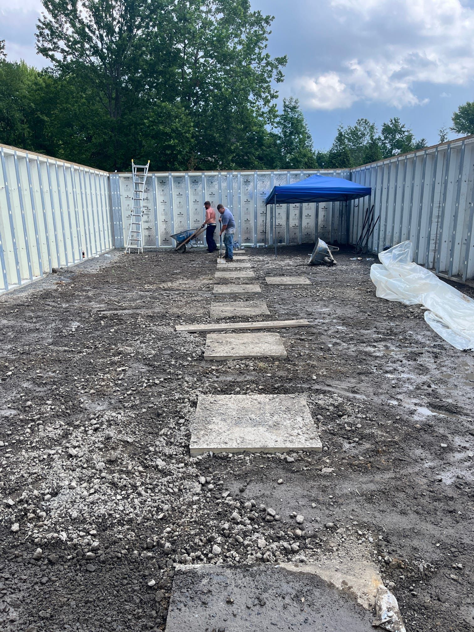 Construction site with workers, gravel, and concrete stepping stones. A blue canopy provides shade.
