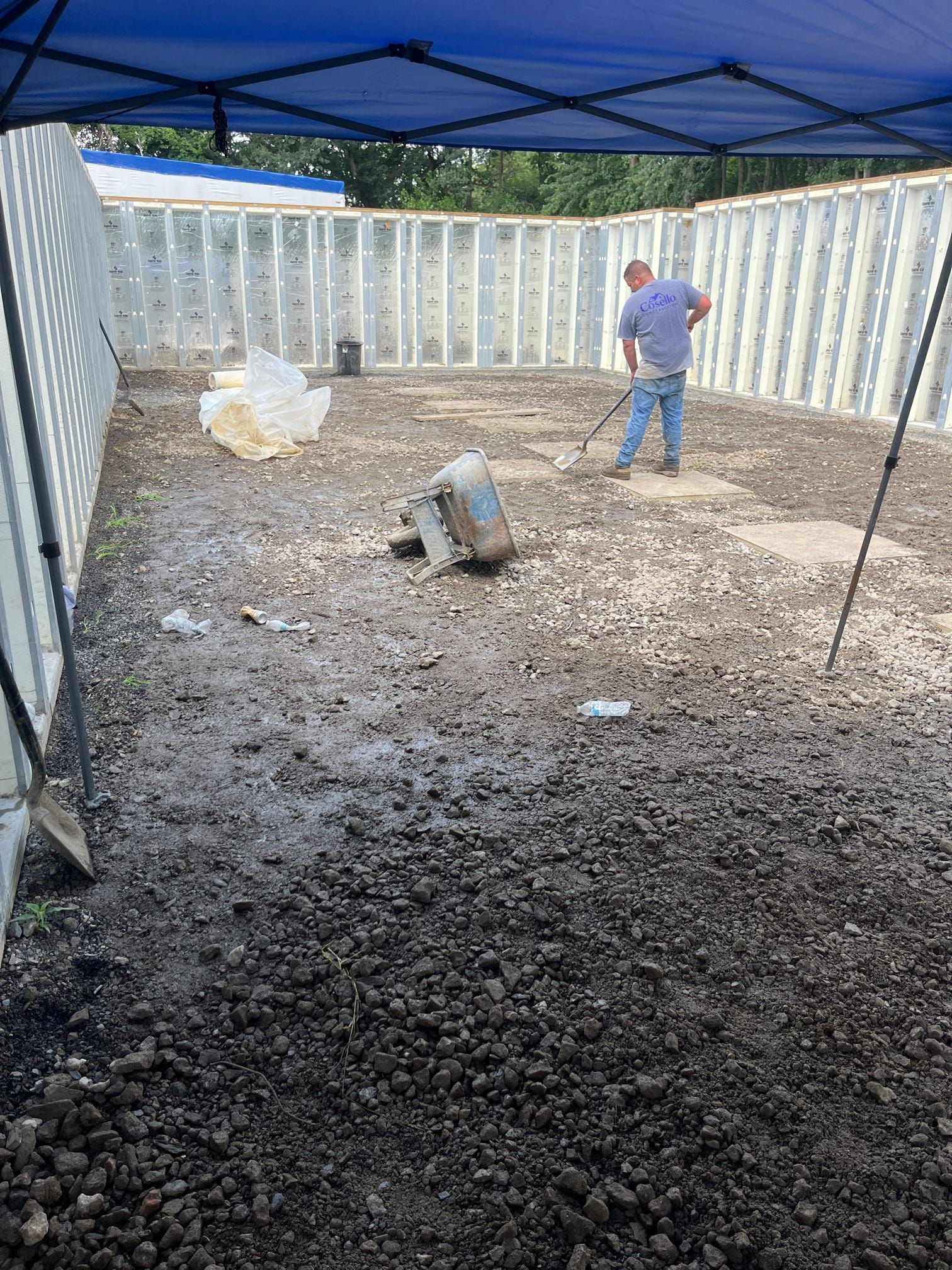 A man rakes a dirt area under a blue canopy; a motor and debris are scattered about.