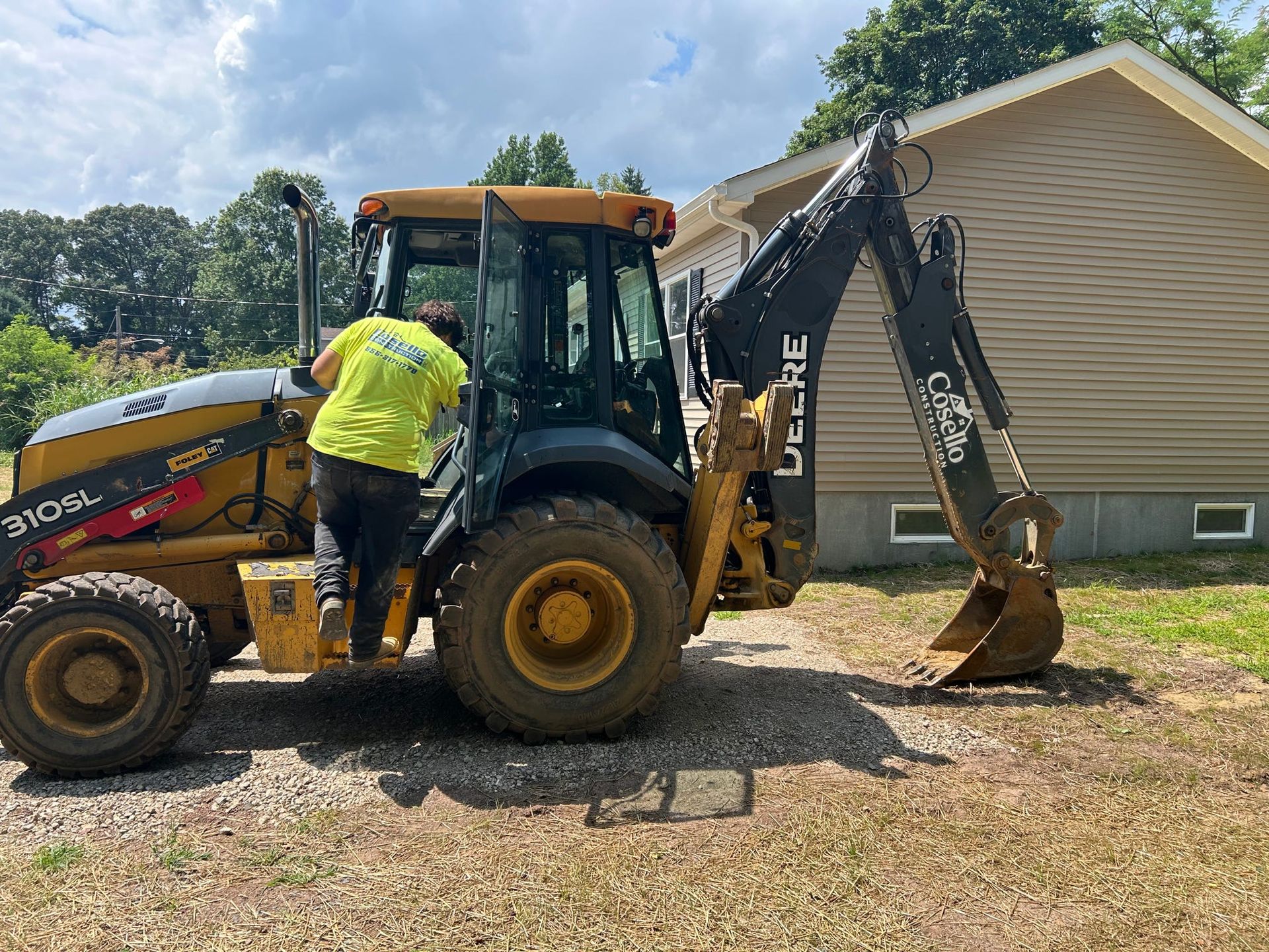 A person in a yellow shirt enters a yellow and black John Deere backhoe in front of a house on a sunny day.