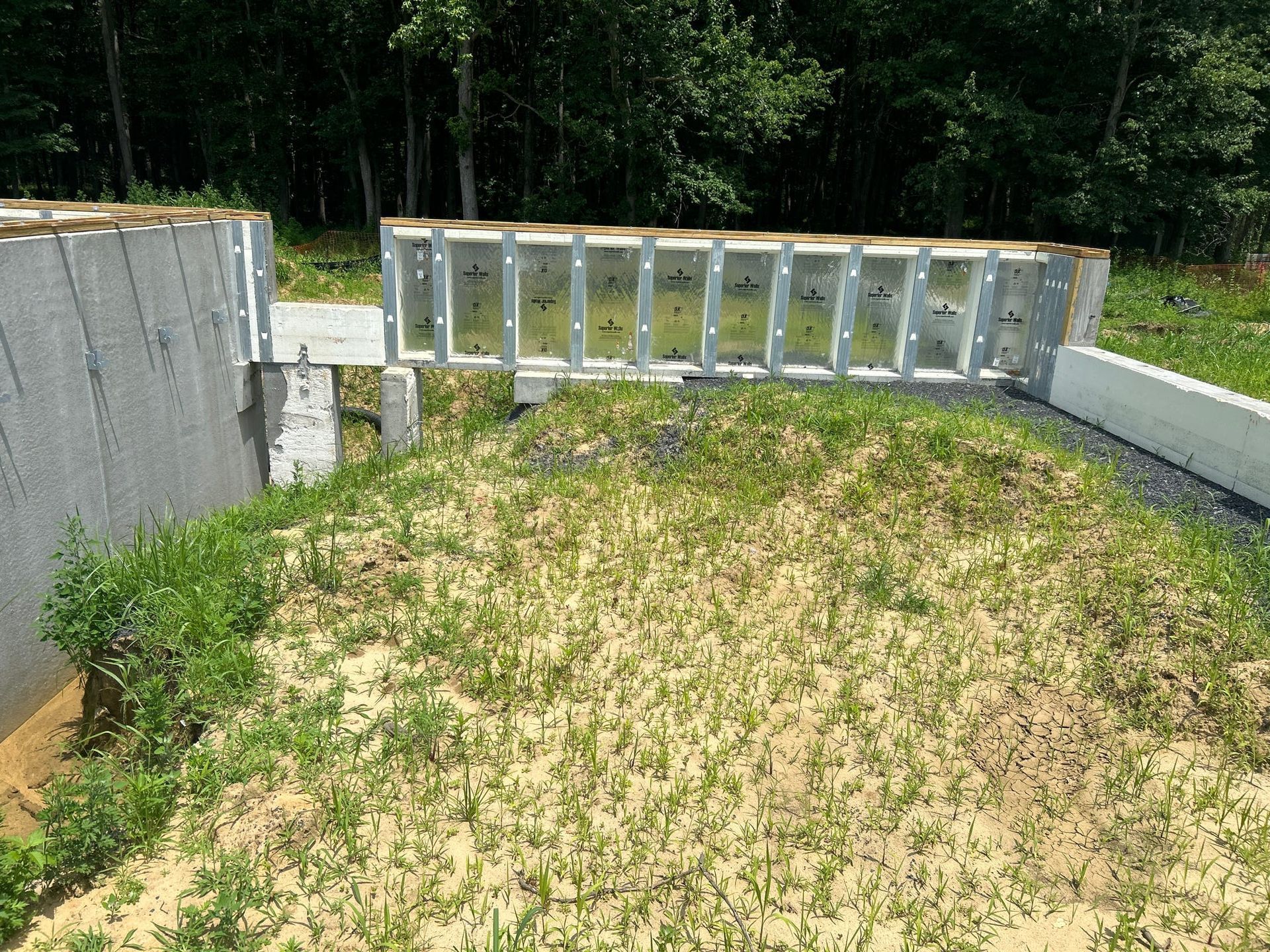 Construction site with gray concrete and metal framing, overgrown with grass and weeds, outdoors.
