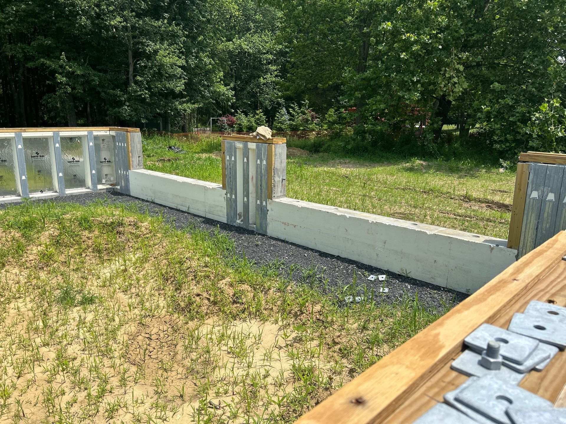 Construction site: concrete foundation walls with metal and wood supports, gravel, and grass.