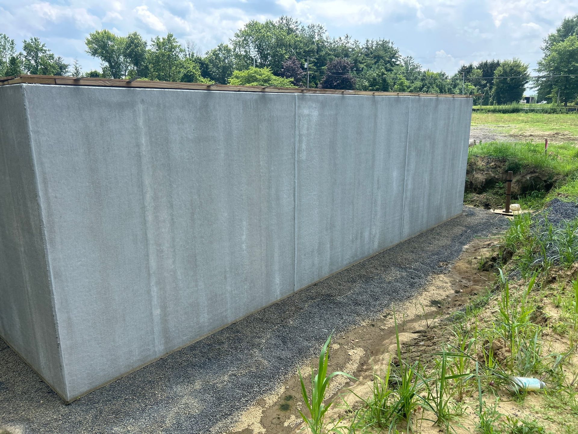Gray concrete retaining wall alongside a gravel path and grassy area, with trees in the background.