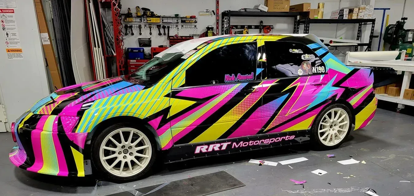 A colorful car wrapped in pink, yellow, and blue with white rims, parked in a garage.
