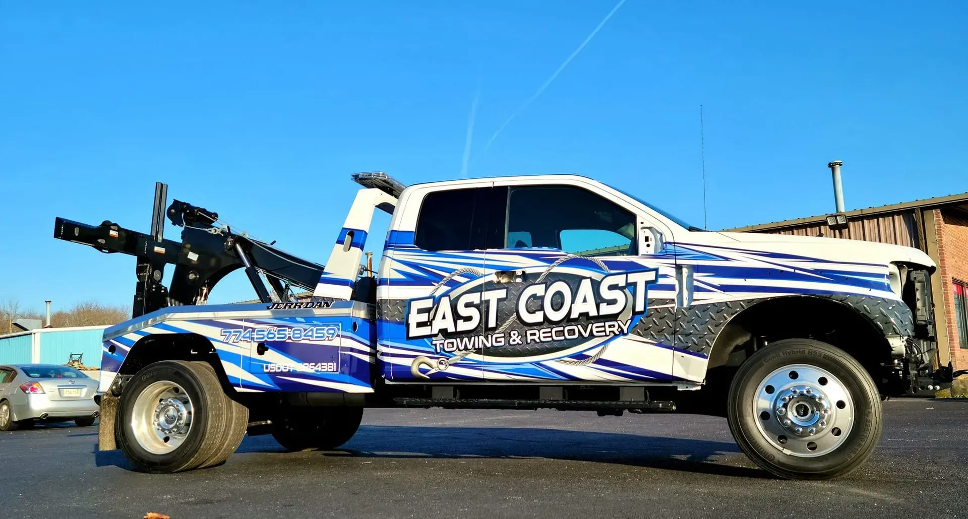 White and blue East Coast Towing truck with boom raised, parked on asphalt.