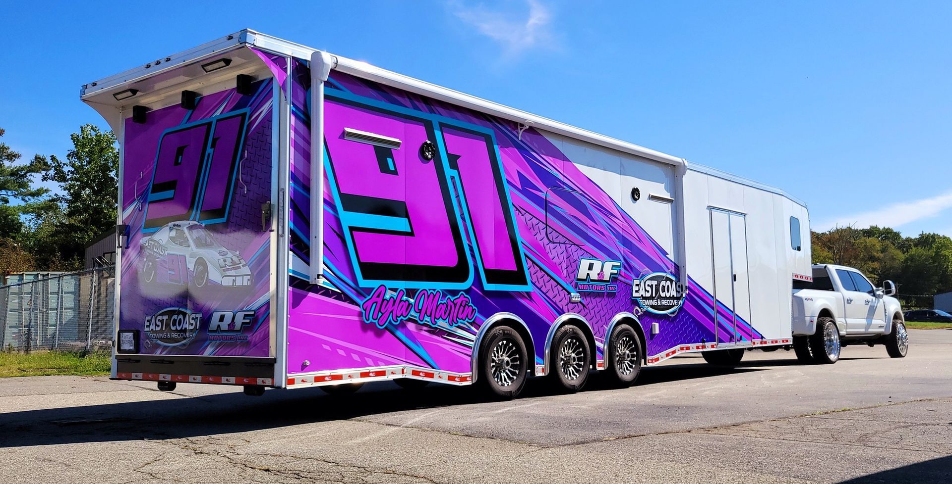 Racing trailer with purple and teal graphics, pulled by a white truck on a sunny day.