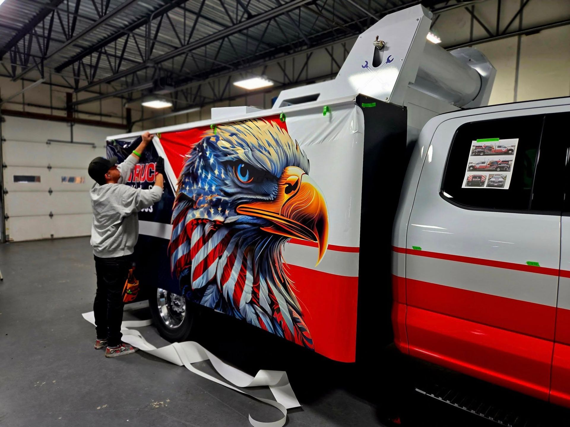 Two people applying a large eagle-themed graphic wrap to the side of a white truck in a garage.
