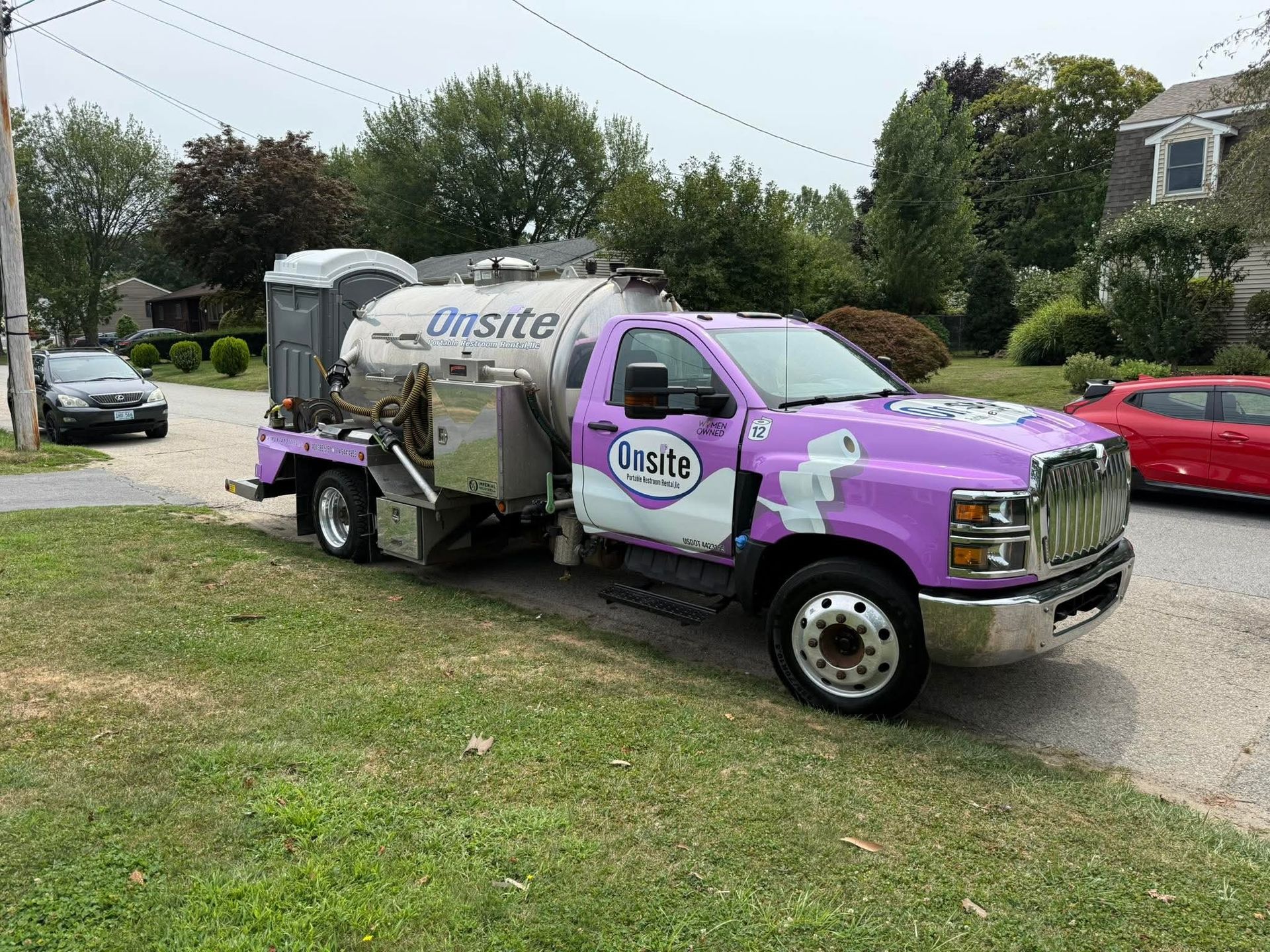 Purple and silver septic truck parked on a residential street with a portable toilet on the back.