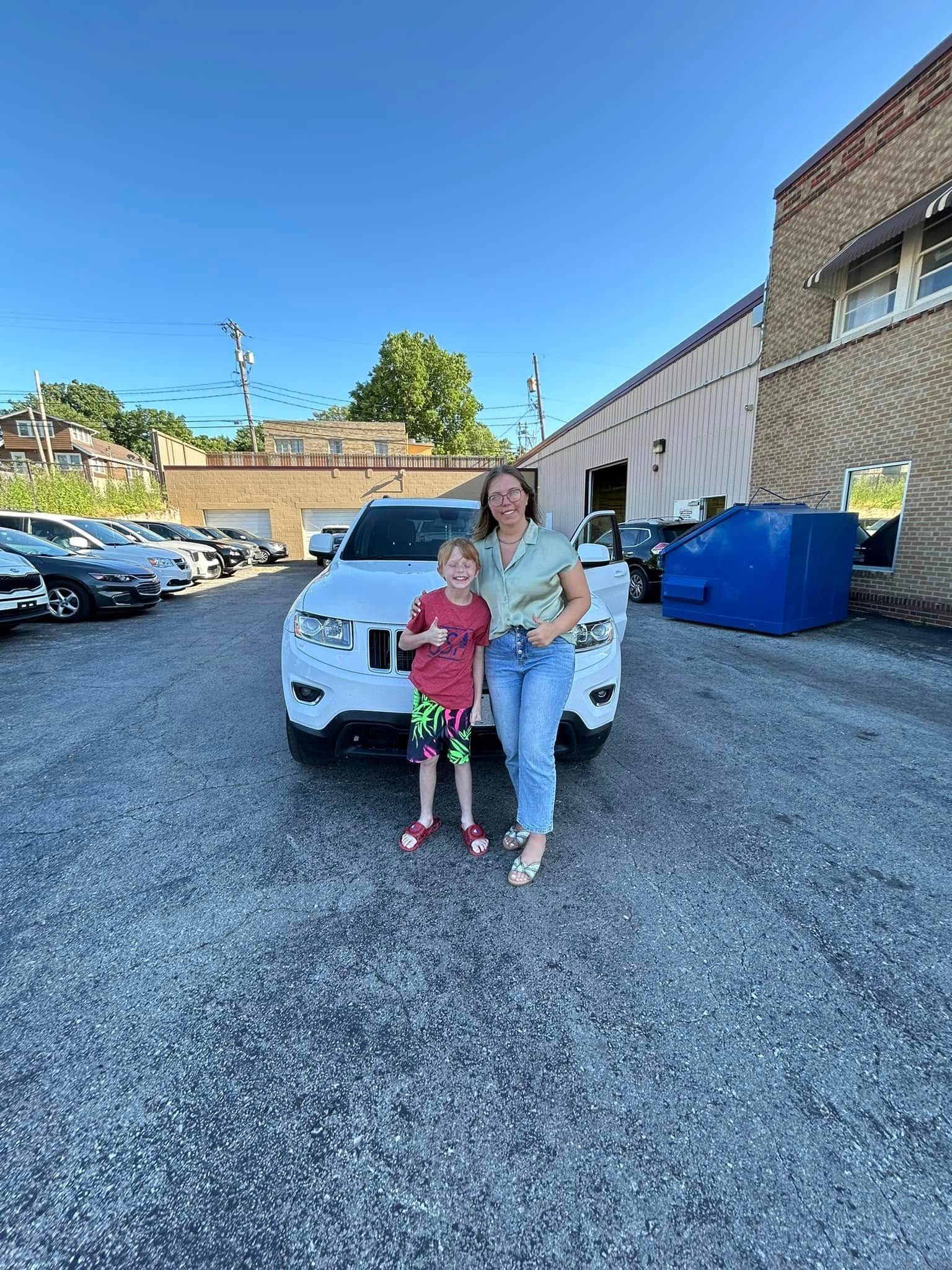 A woman and a child are standing in front of a car in a parking lot.