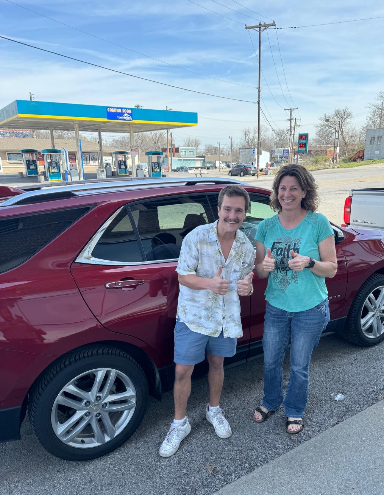 A man and a woman are standing in front of a red car giving a thumbs up.