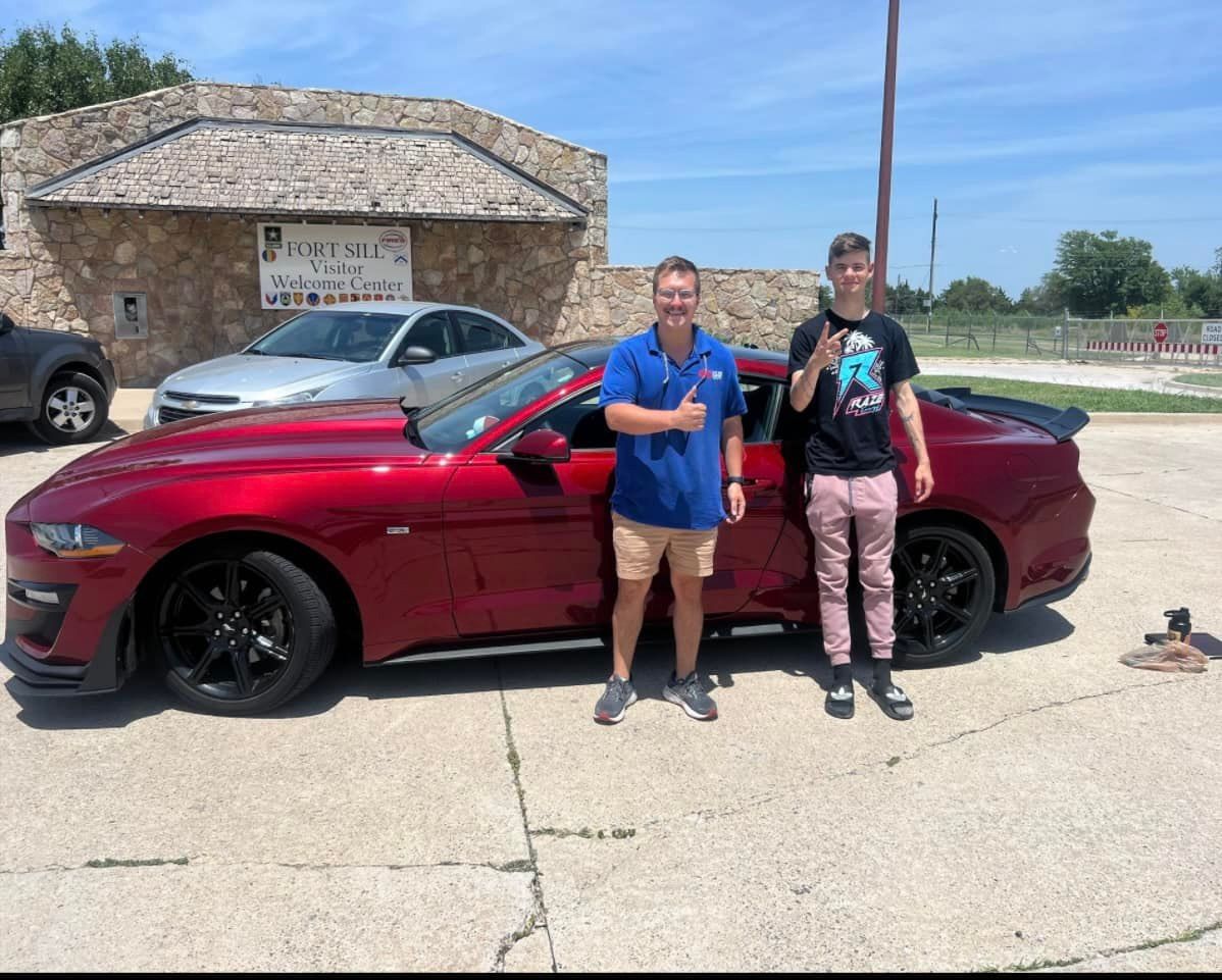 Two men are standing in front of a red car.