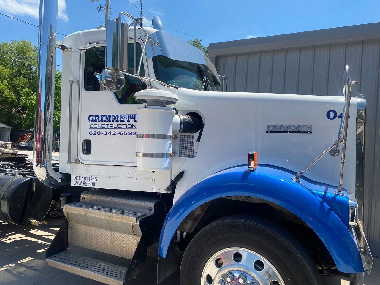 White and blue semi-truck with chrome details parked outside a grey building; 'Grimmett' is printed on the door.