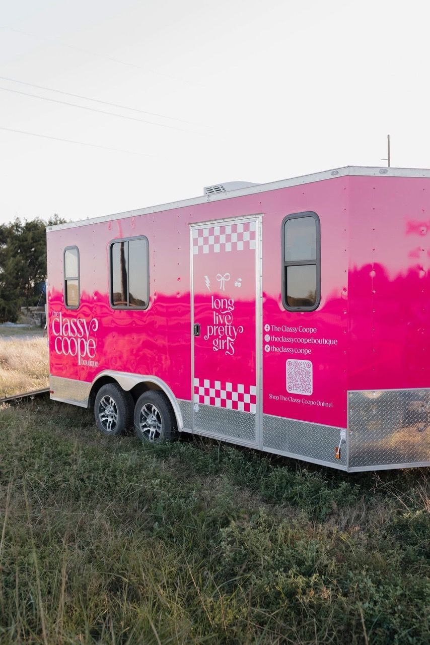Pink food trailer with checkered door and windows, parked on grassy field.