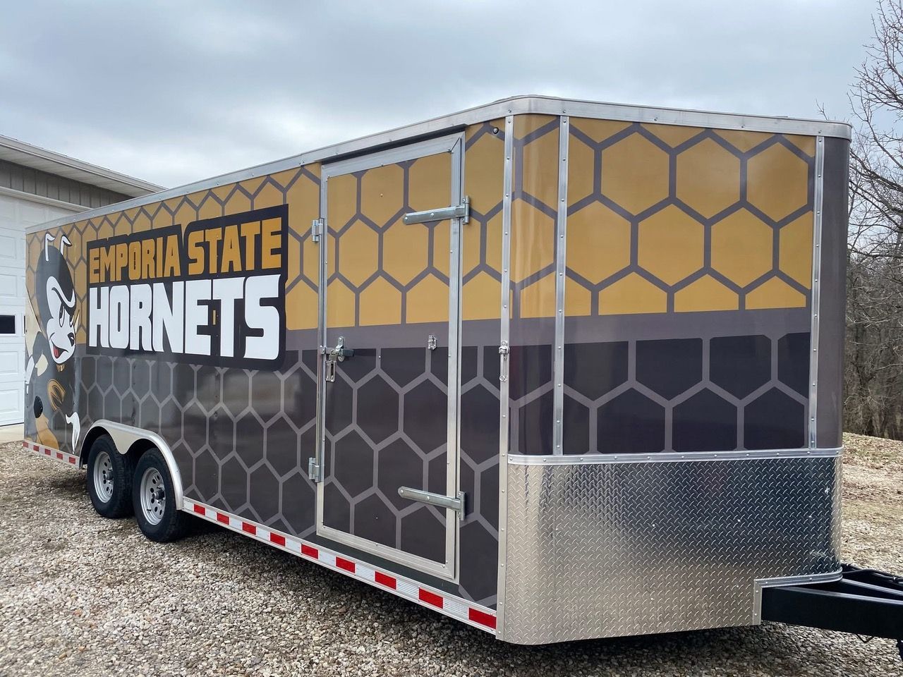 Enclosed trailer with Emporia State Hornets logo and honeycomb pattern, parked outdoors on gravel.