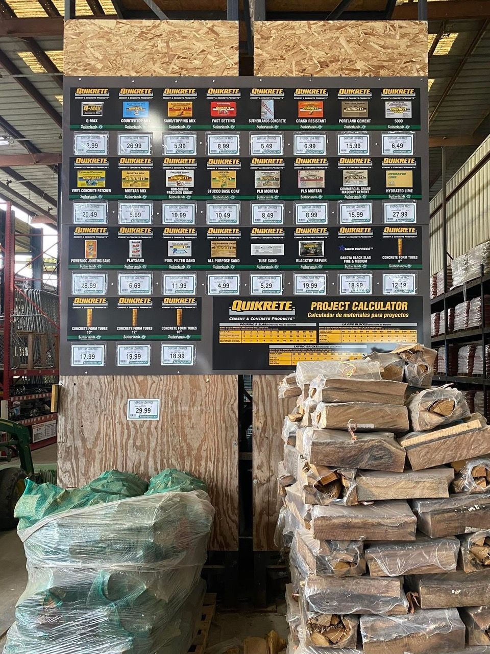 Sign displaying firewood types and prices next to a stack of firewood and a bag of wood chips.