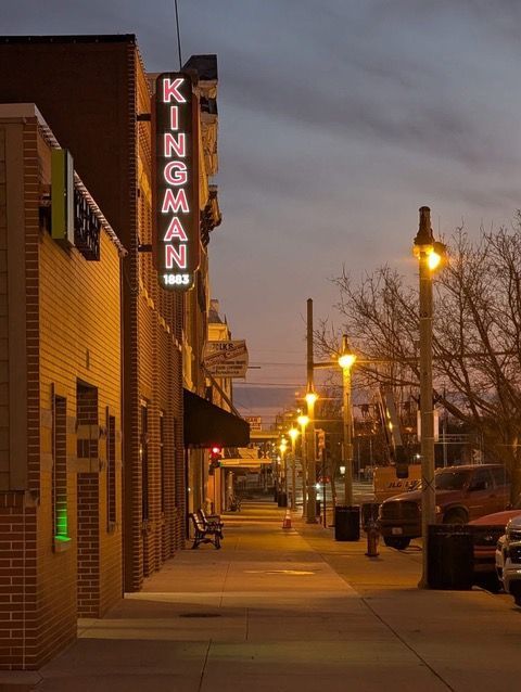 Kingman neon sign on brick building at dusk, street with streetlights and parked vehicles.